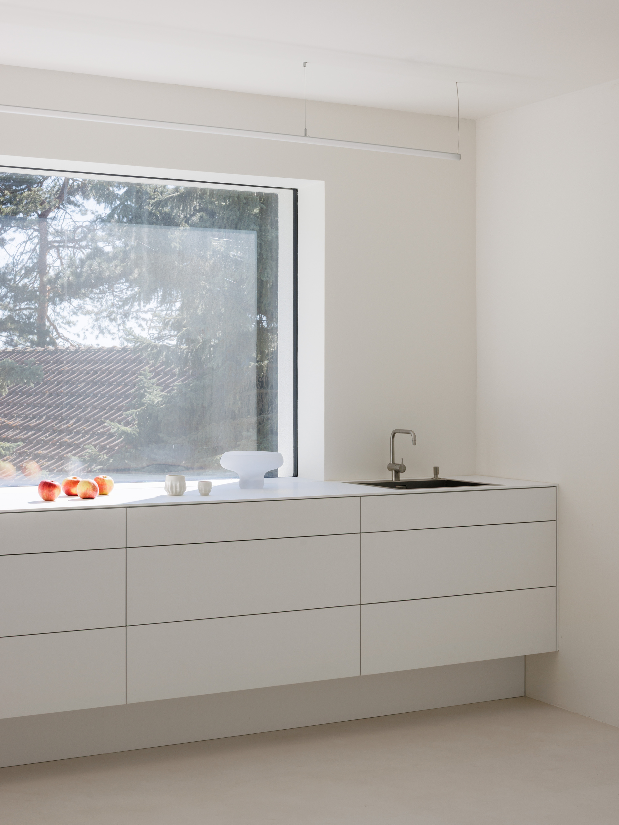 White handleless kitchen cabinetry beneath a horizontal window overlooking pine trees in soft light