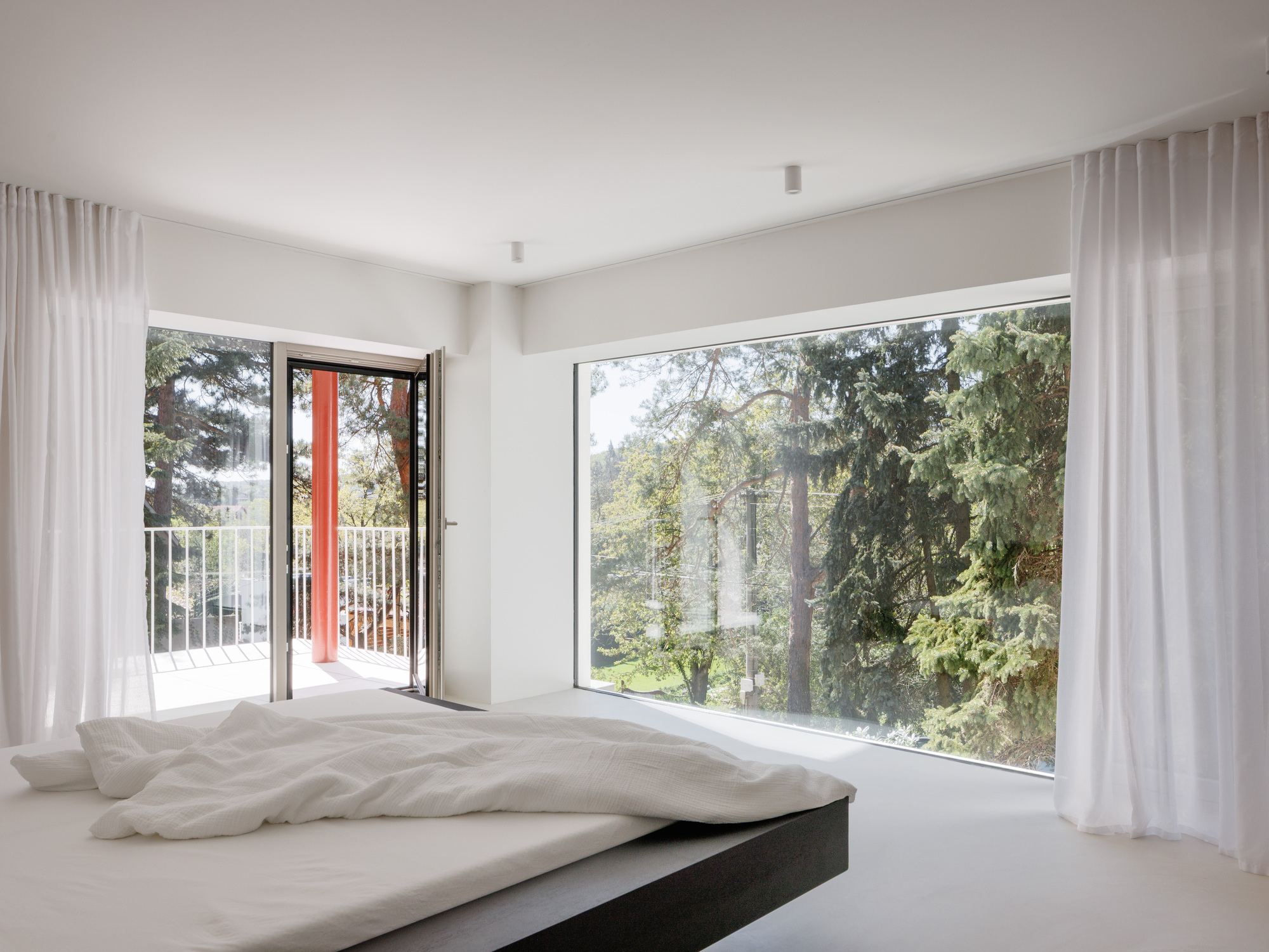 Corner bedroom windows framing treetops with sheer drapes and an open balcony door