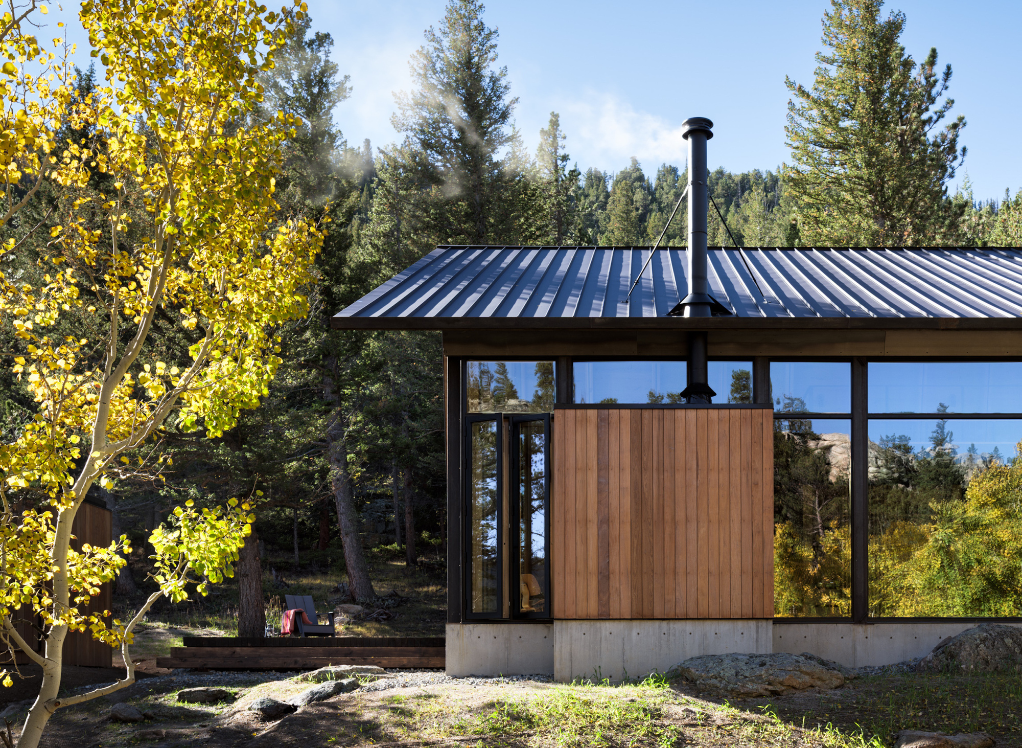 Exterior view showing standing seam metal roof, timber and glass facade, and concrete plinth among aspens