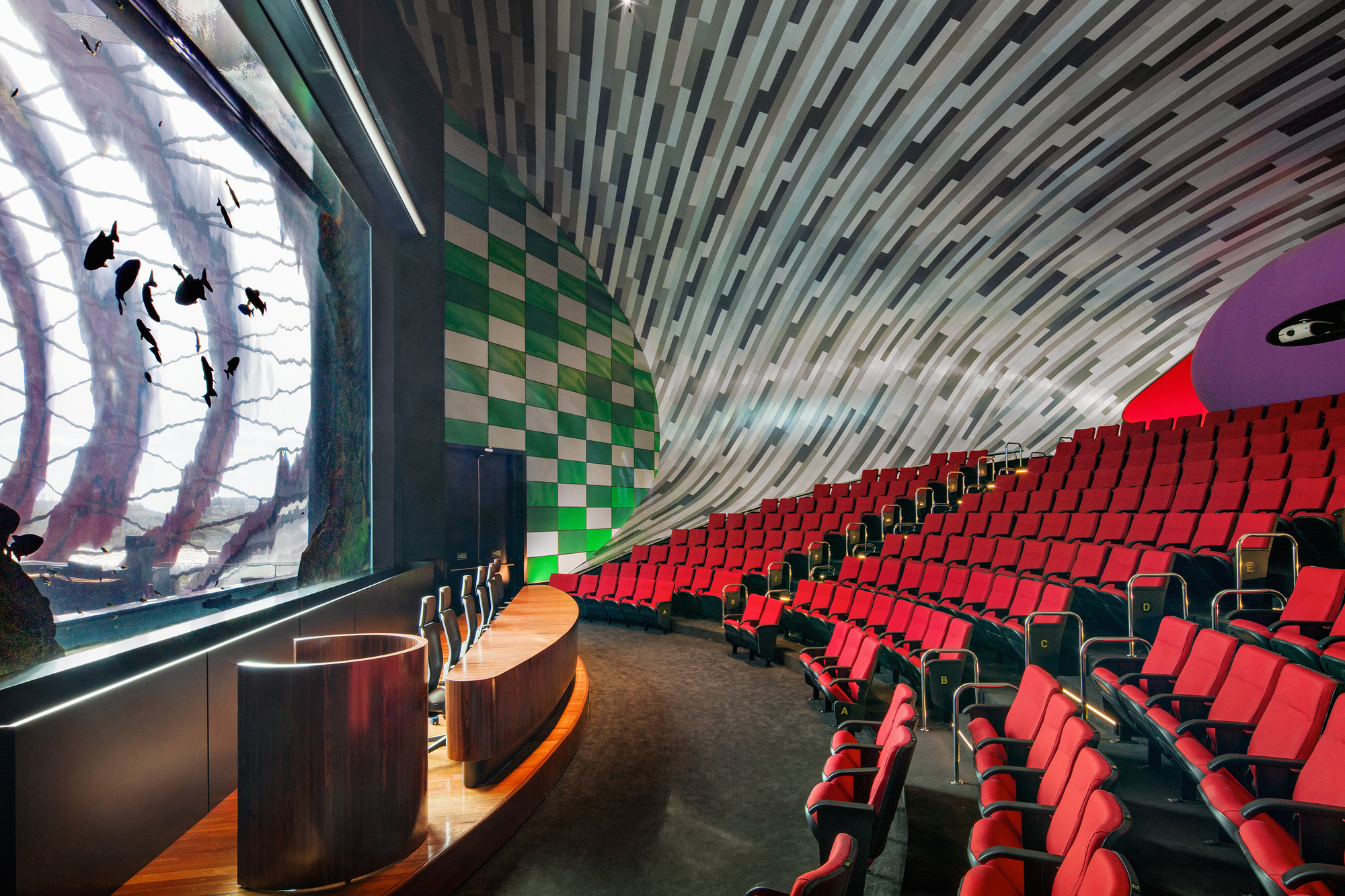 Auditorium interior with red seating rows and ribbed ceiling beneath a window wall with silhouette artwork
