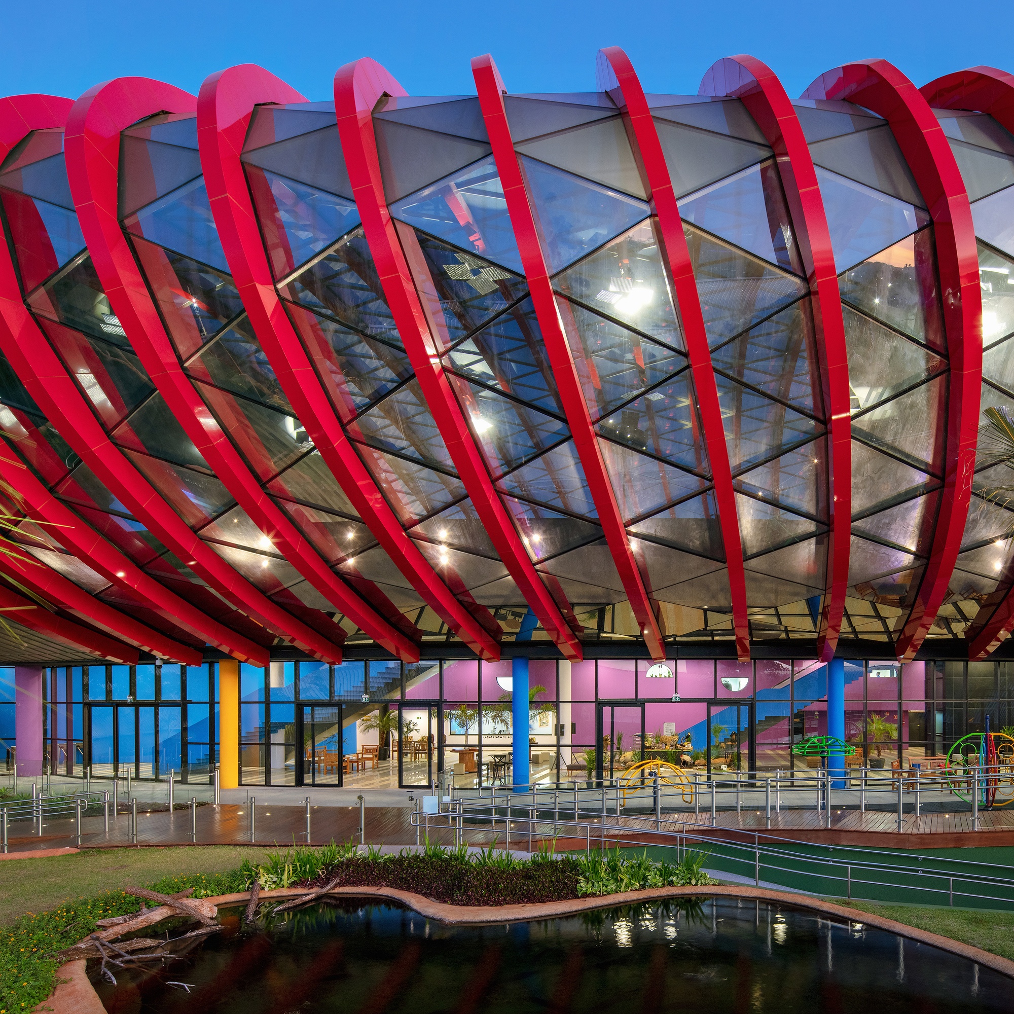 Exterior facade with red arched ribs wrapping glazed envelope above a reflecting pond at twilight