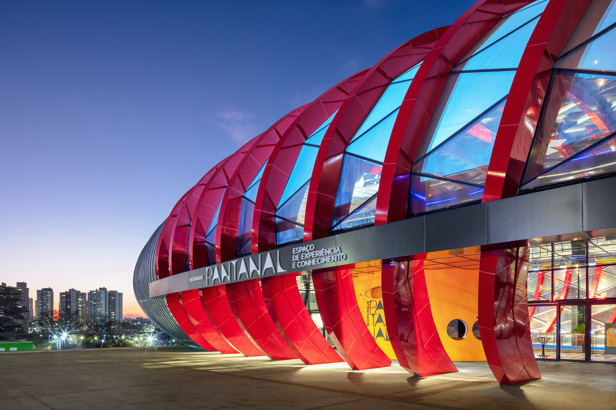 Entry canopy showing red structural ribs arching over glass walls at dusk with warm interior lighting