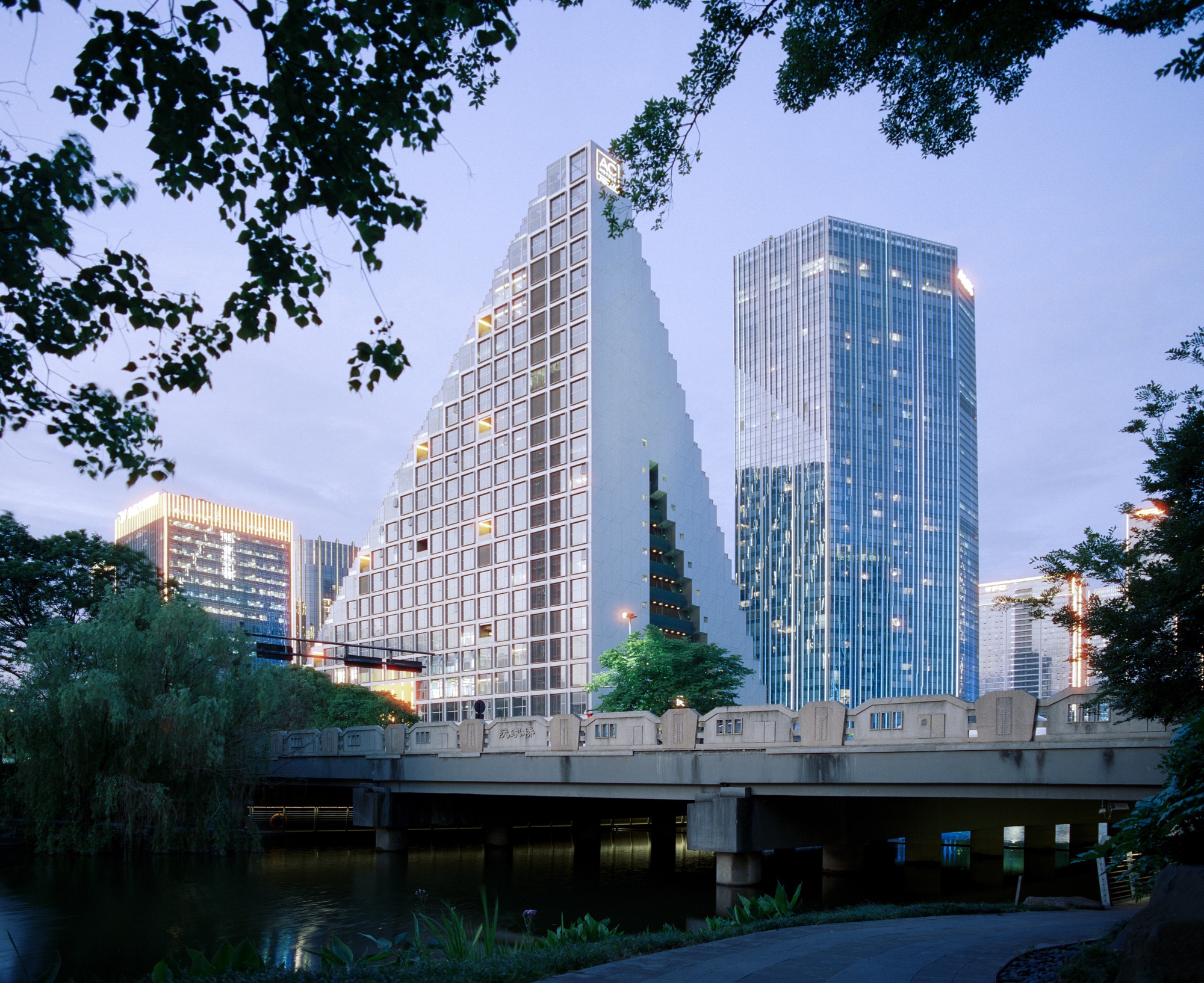 Stepped tower with pixelated facade framed by tree branches and viewed across the river at dusk