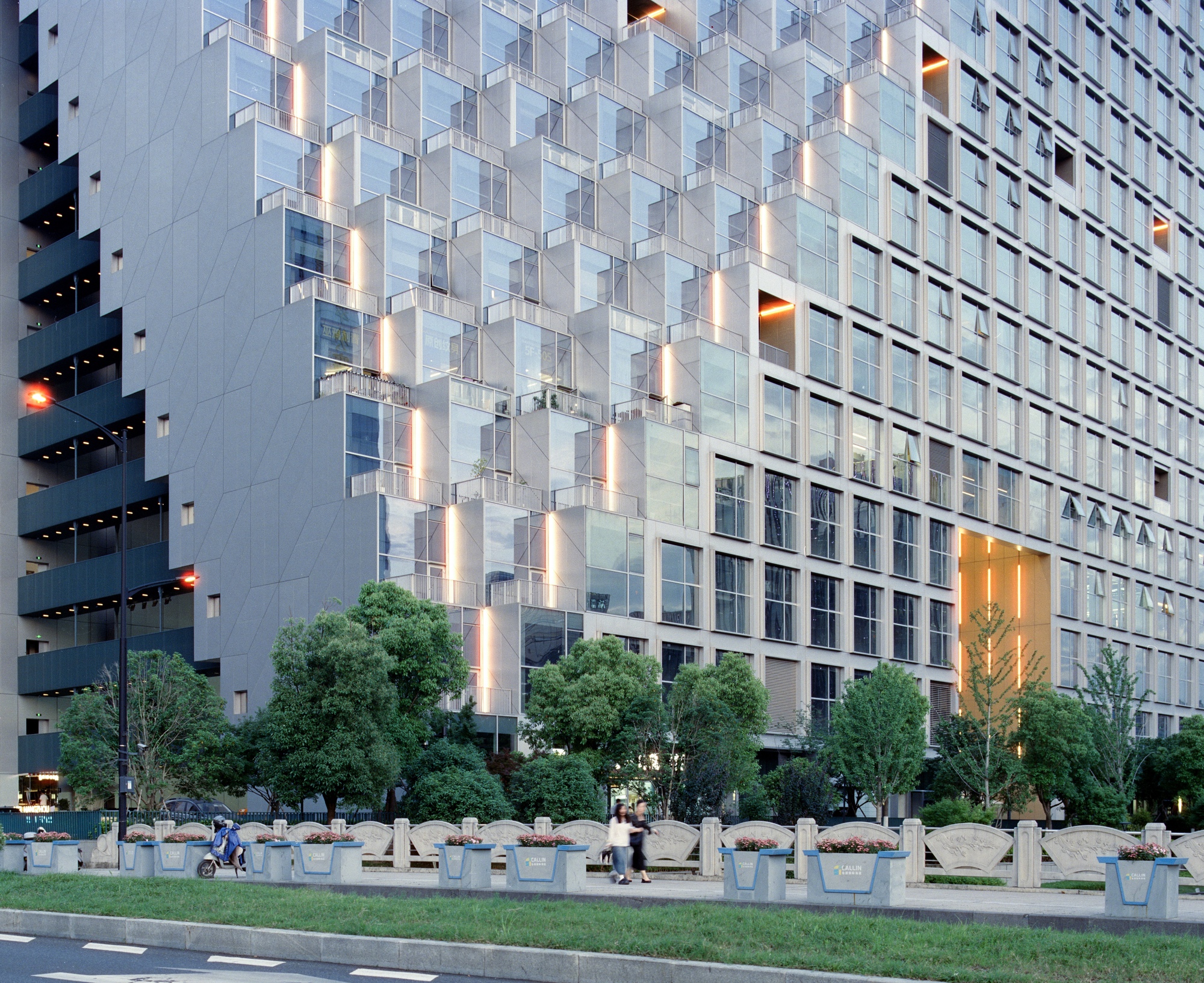 Ground-level view showing the modular facade above a row of planters and pedestrians at twilight