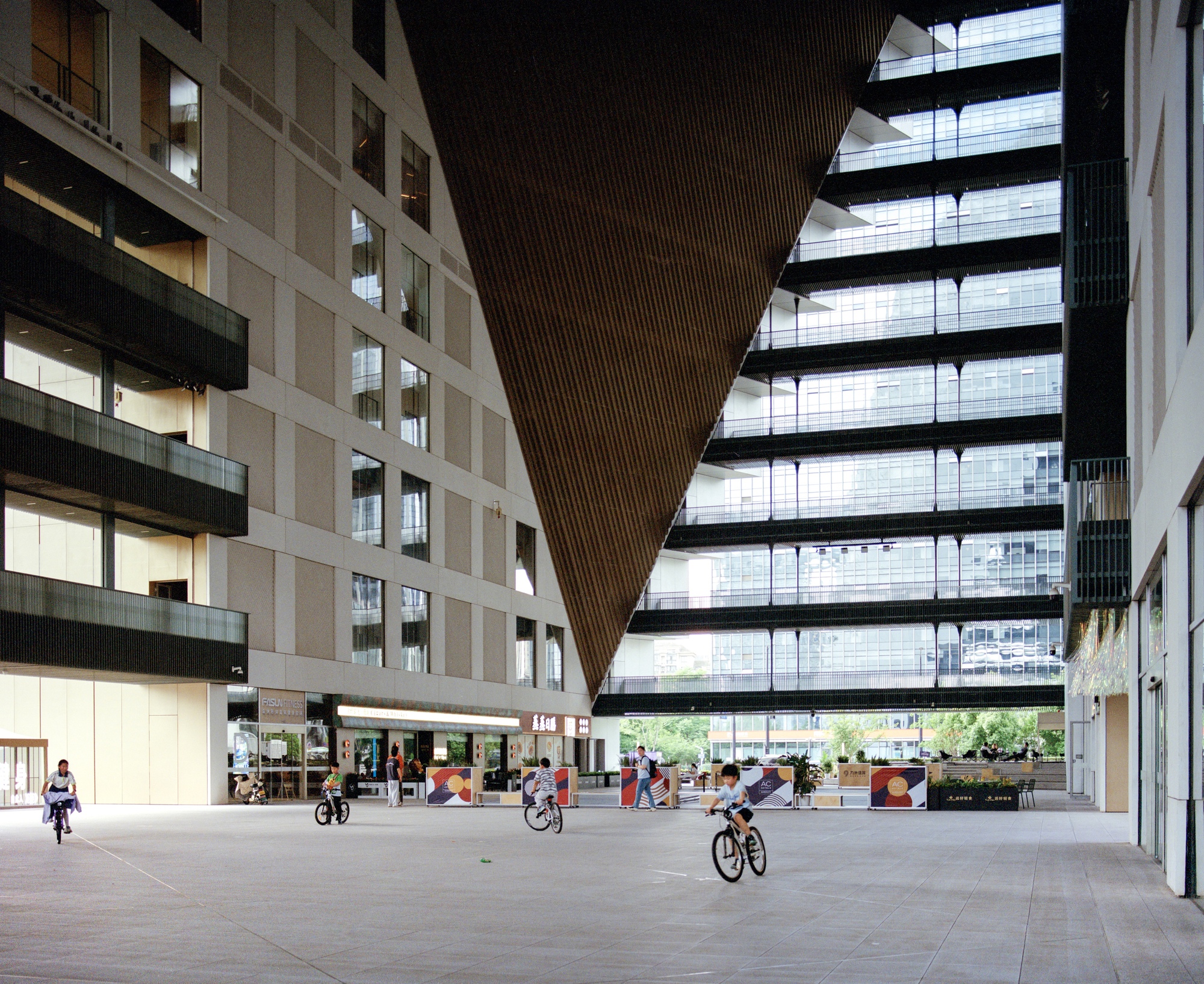 Covered courtyard beneath a triangular slatted ceiling with cyclists passing through the open plaza