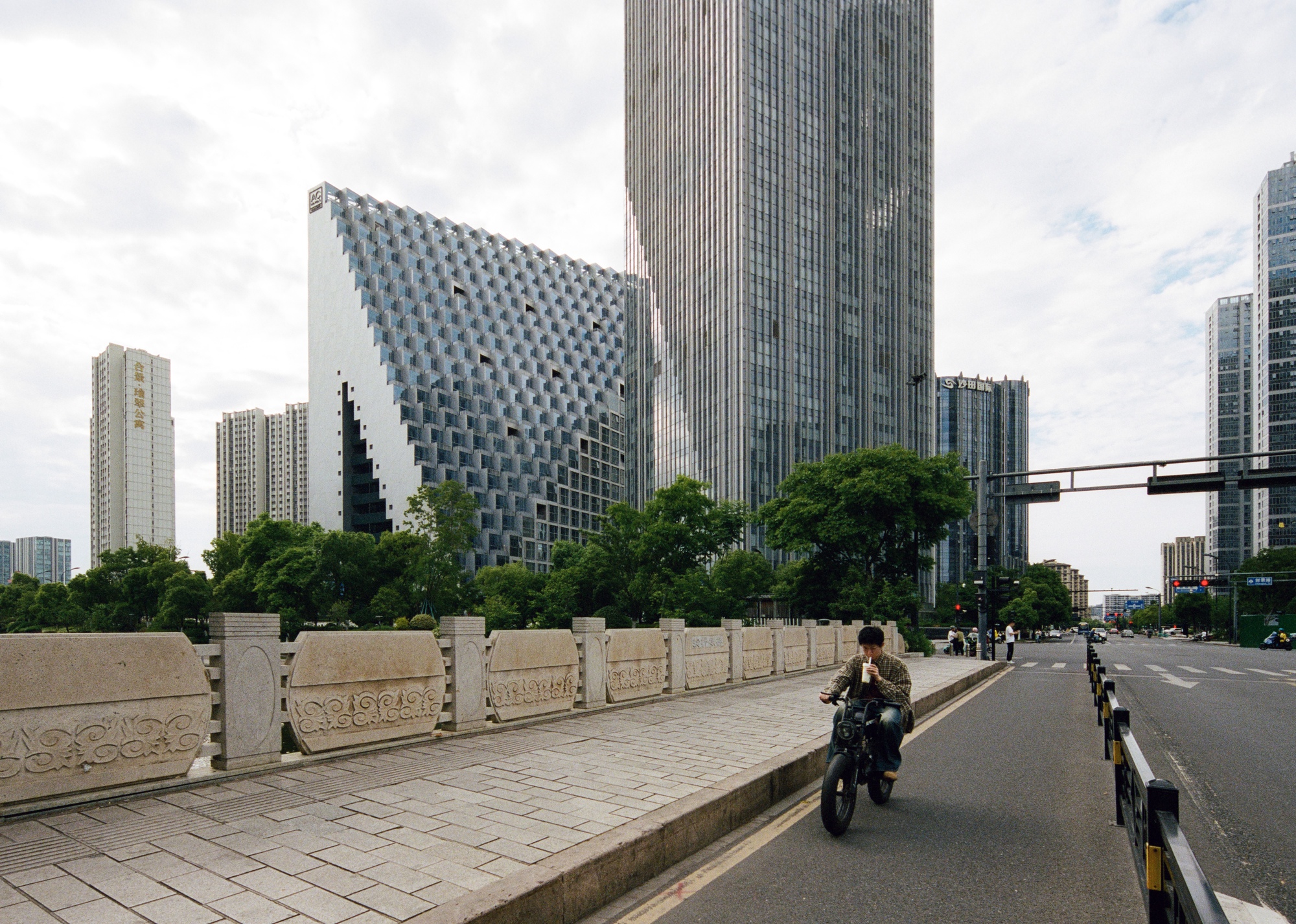 Street view of the angled tower with a motorcyclist riding along the tree-lined boulevard