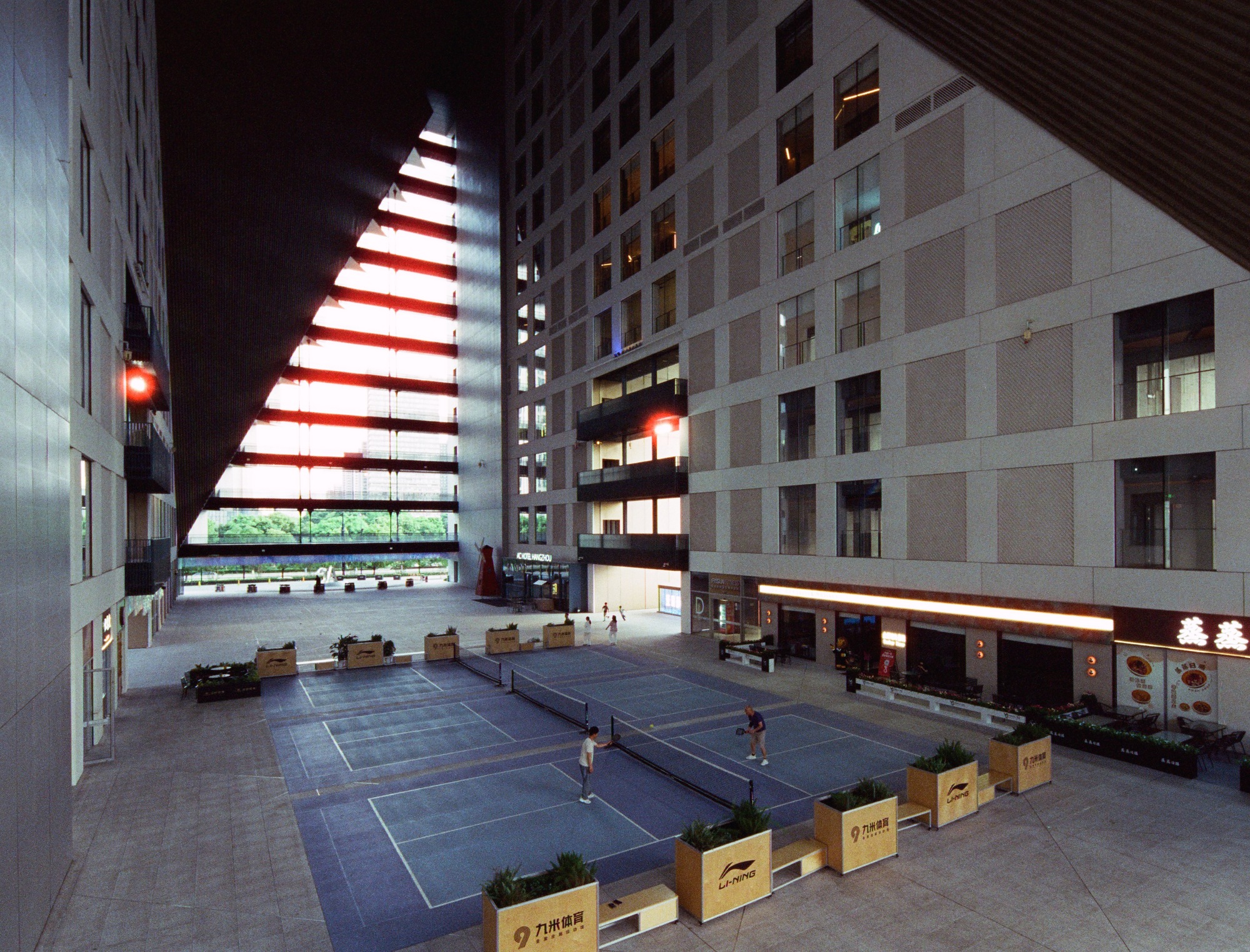 Nighttime courtyard with illuminated triangular opening and wooden seating boxes on the paved plaza