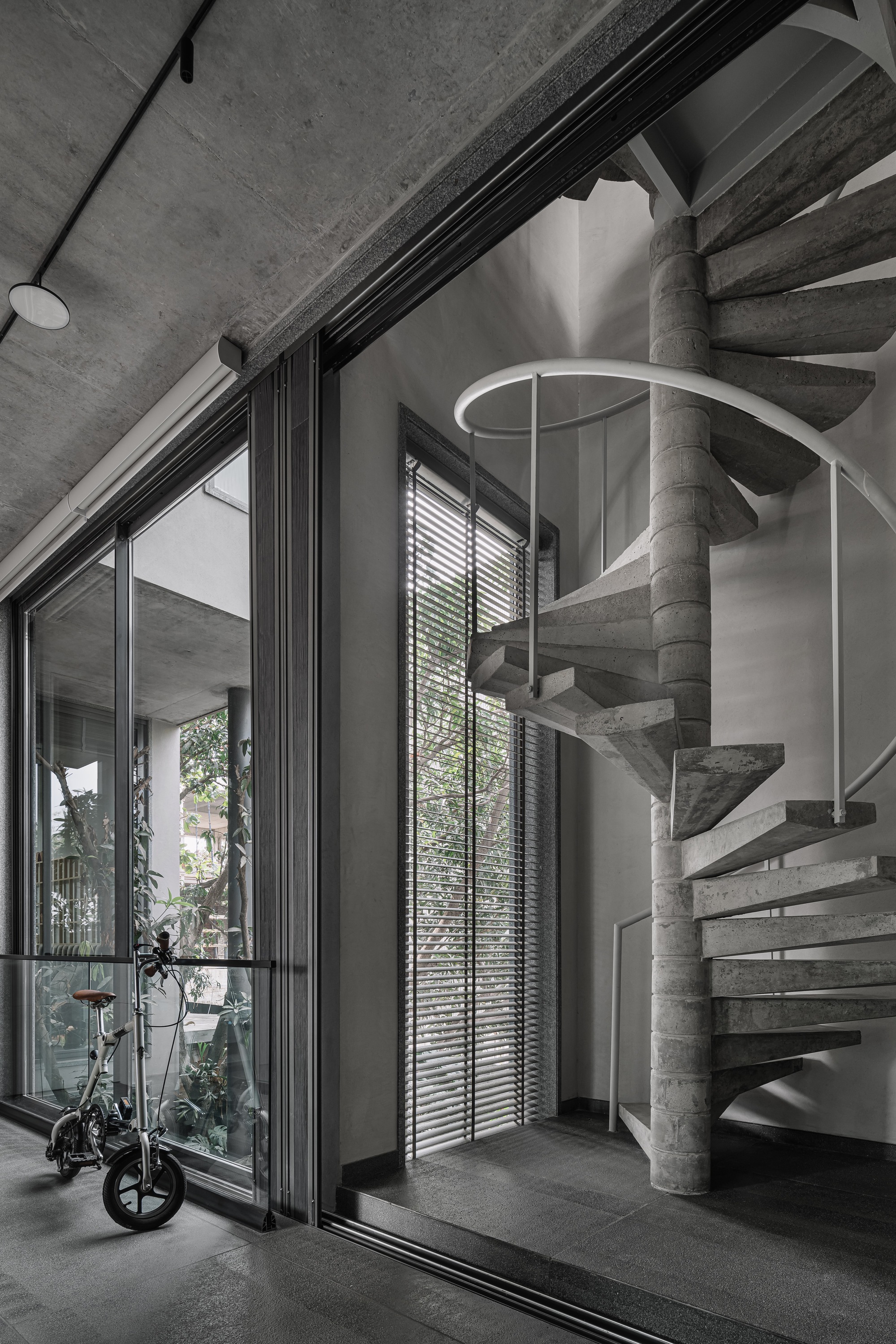 Helical concrete staircase with white metal railing beside floor-to-ceiling glazing and horizontal blinds