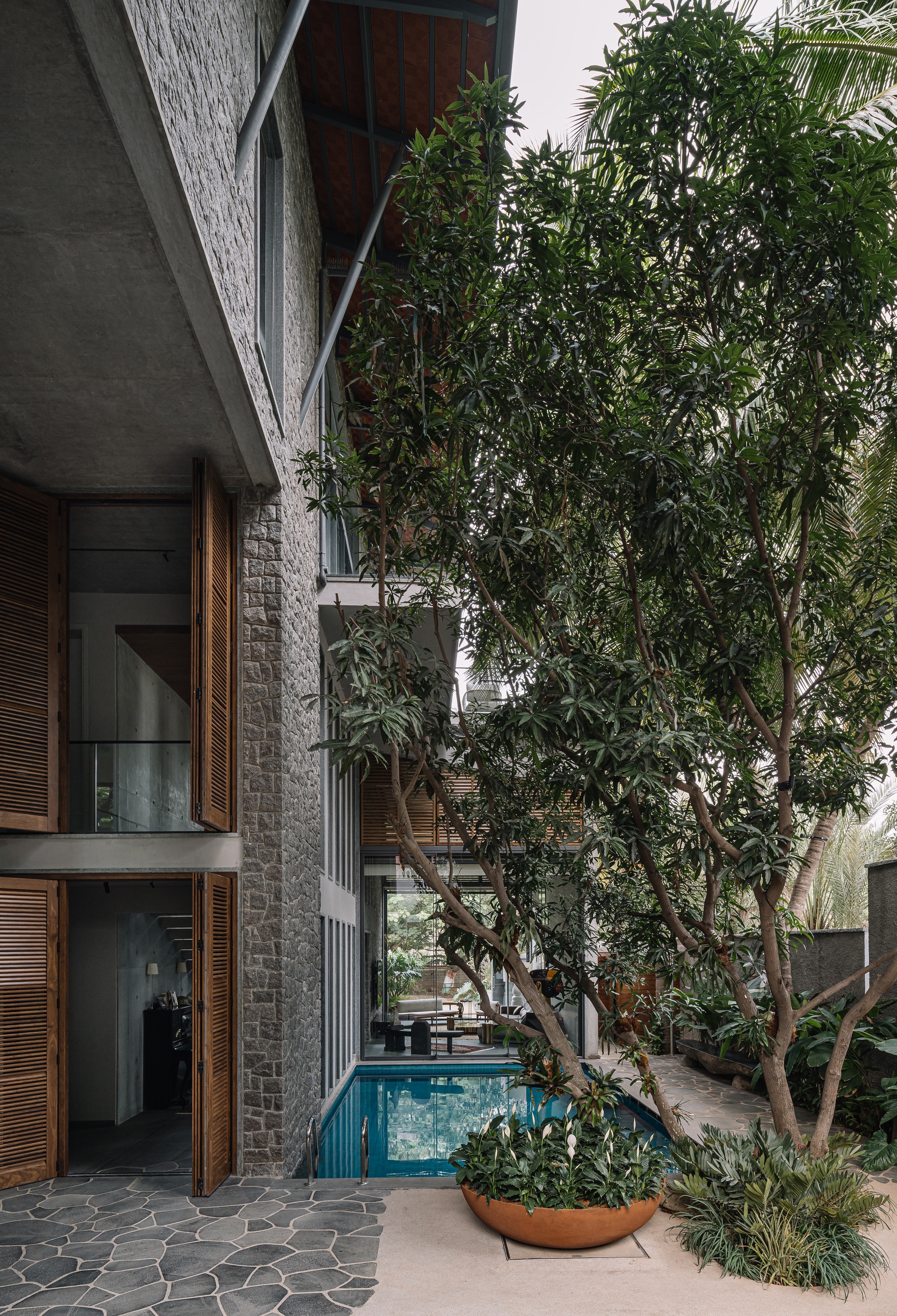 Exterior corridor with textured stone wall, timber louvered doors, and pool beside mature courtyard trees