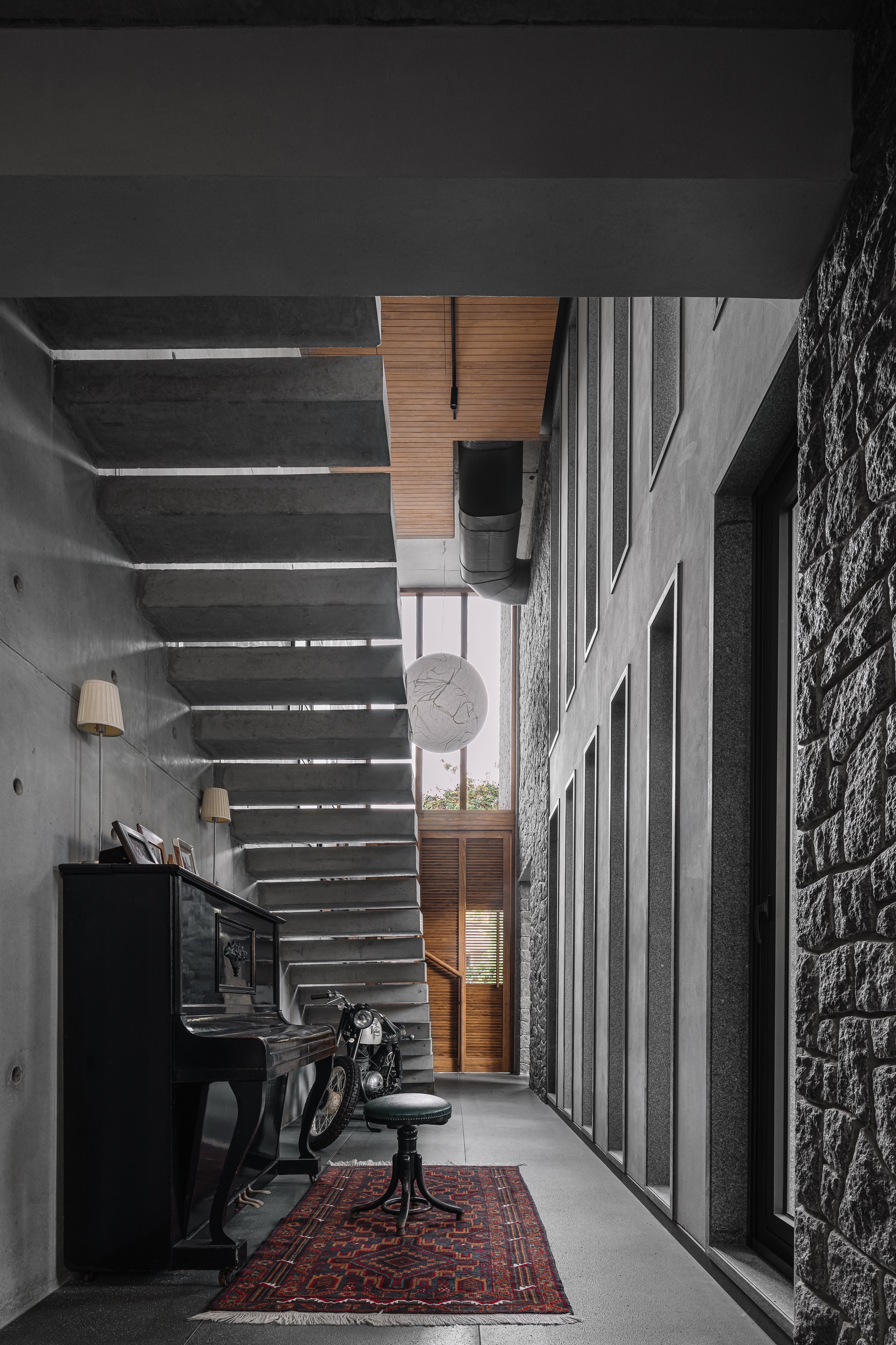 Interior corridor with upright piano, exposed concrete ceiling, horizontal slatted screen and stone wall under natural light