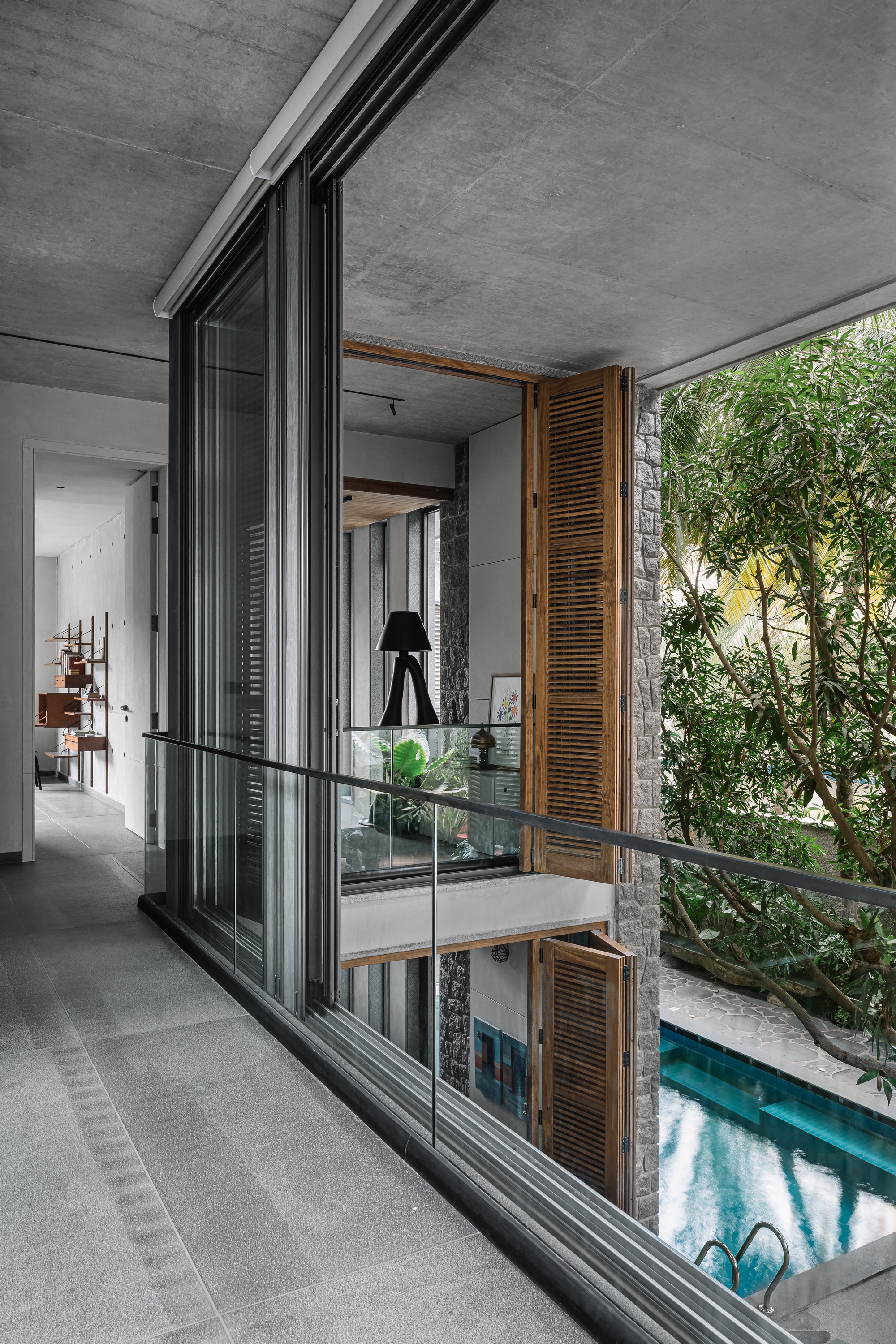 Upper-floor corridor with full-height glazing, timber louvered shutters, and view down to interior pool and greenery