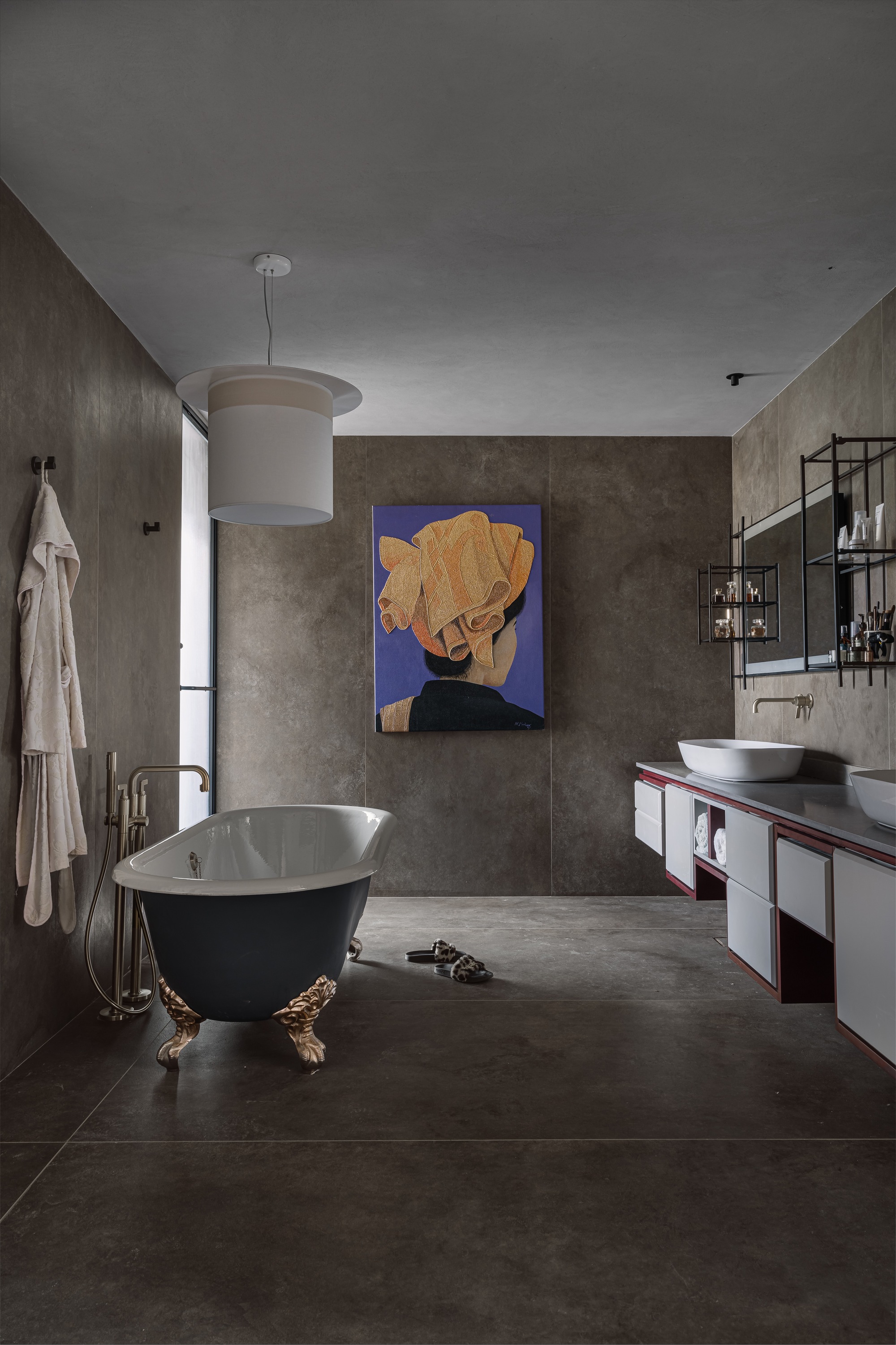 Bathroom with freestanding black tub on brass feet, concrete walls, and abstract portrait painting above