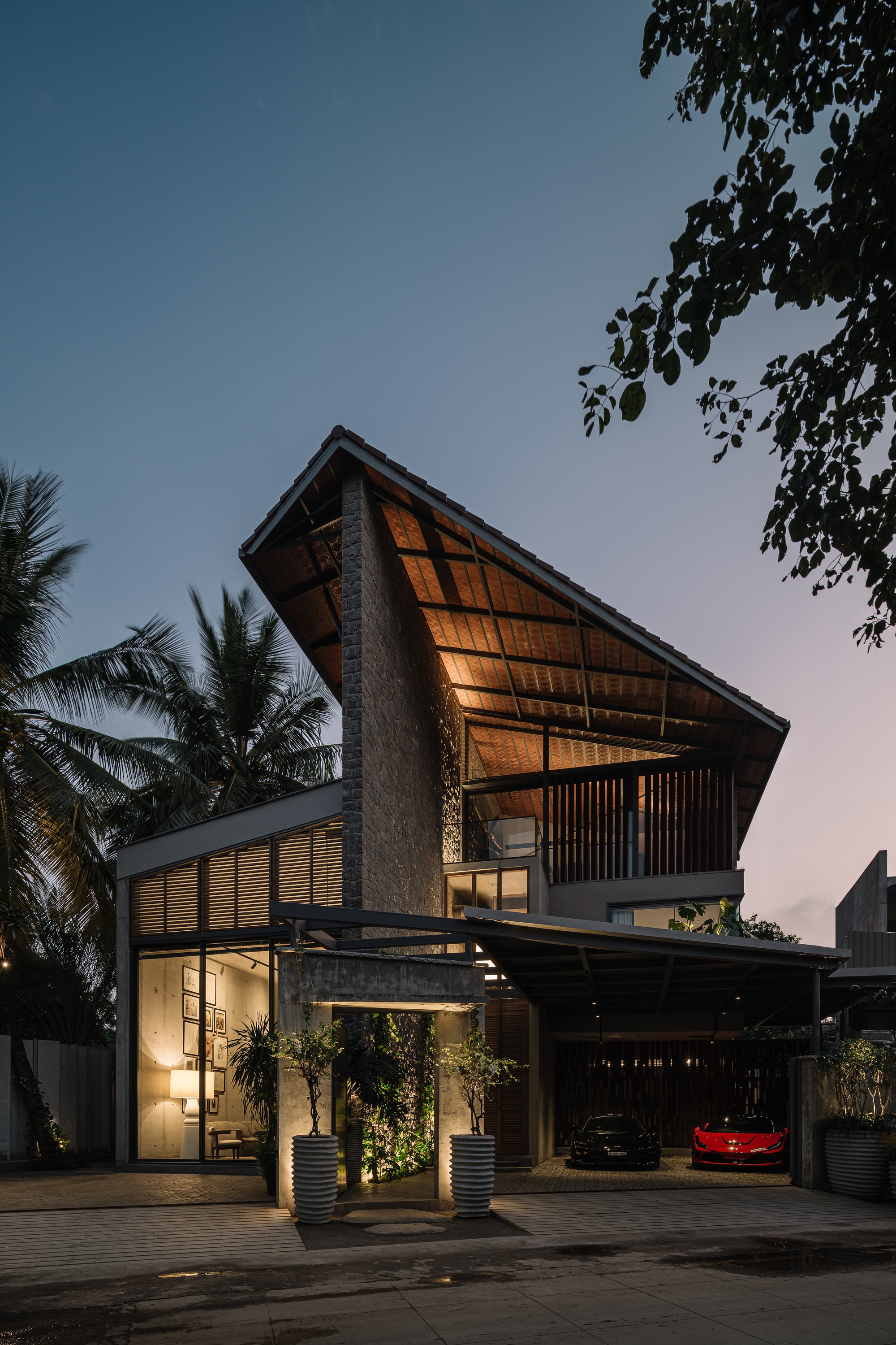 Street view of residence with angled glazed roof and stone facade at dusk beneath palm trees