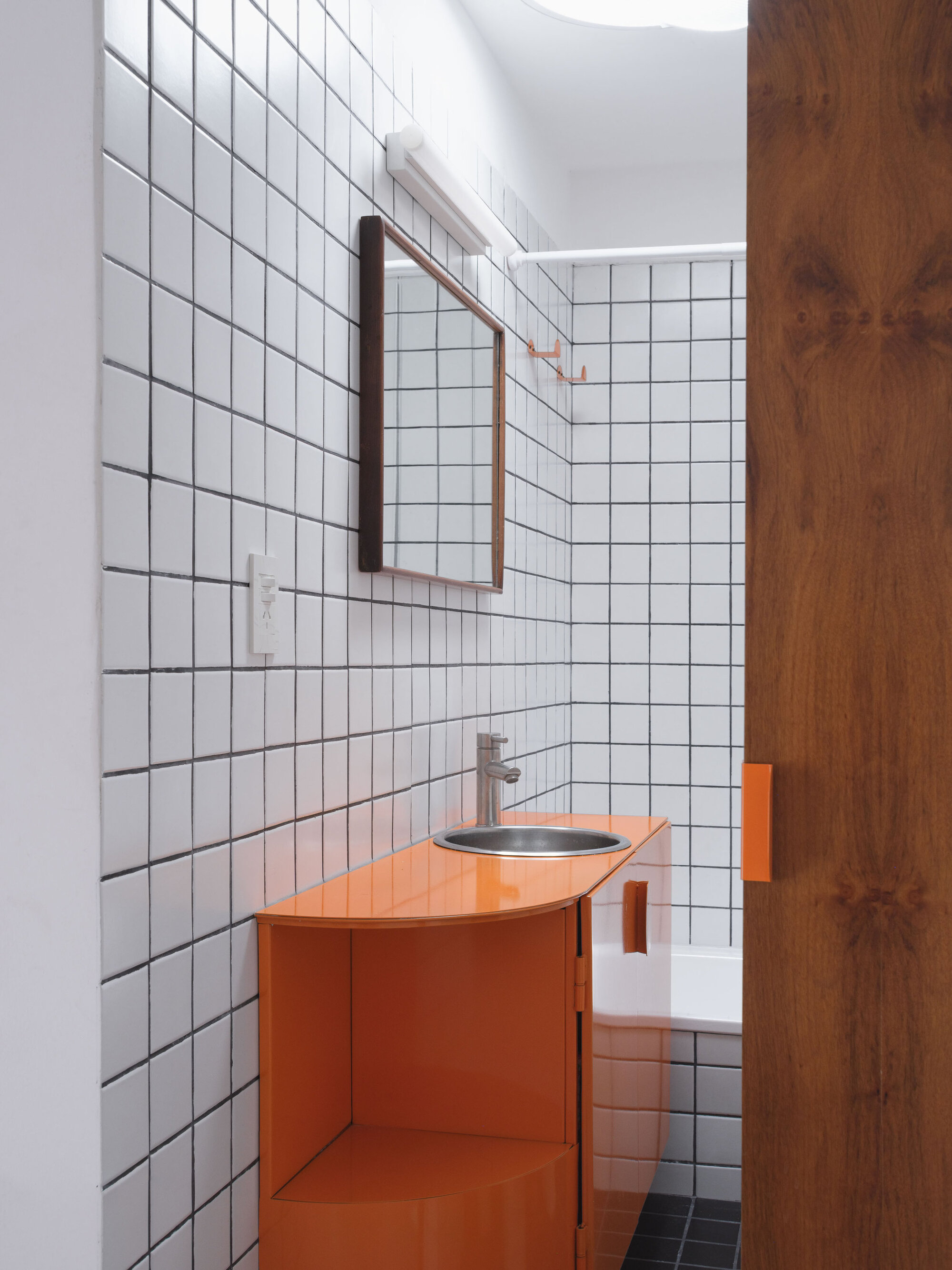 Orange vanity unit with stainless steel basin against white square tile walls beside a timber door