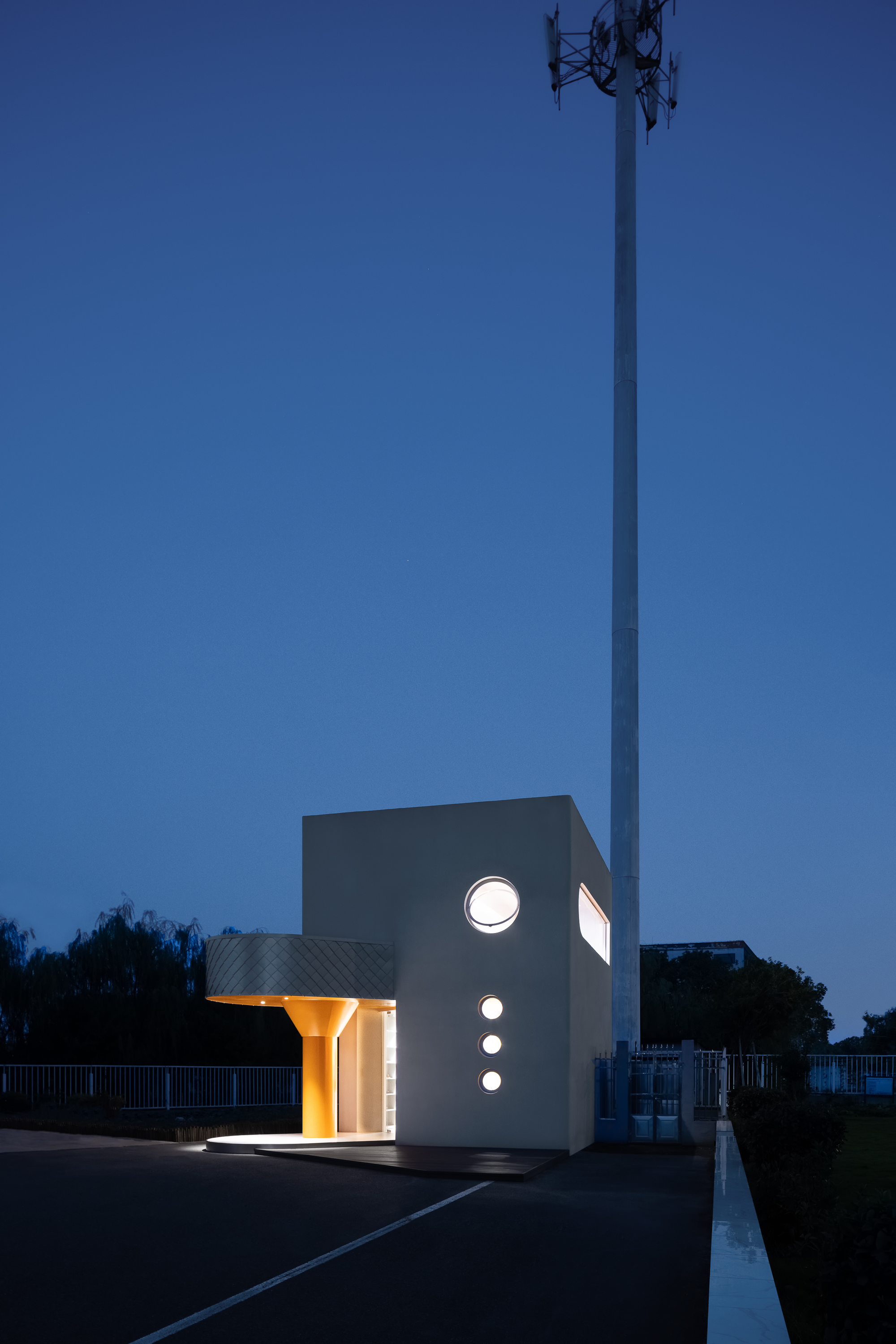 White cubic pavilion with circular portal windows and orange column lit at dusk beside a communications tower