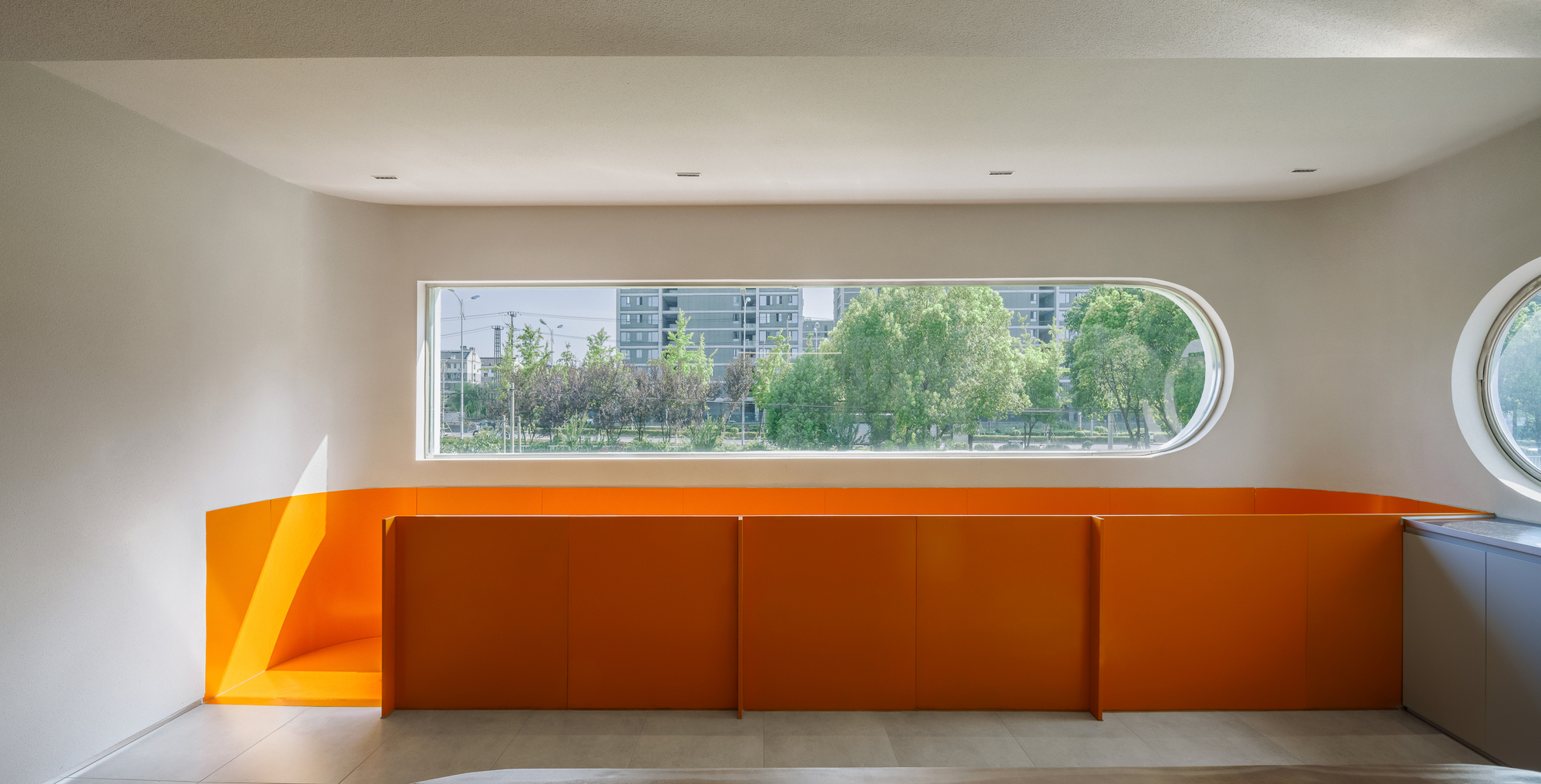 Interior with orange lacquered cabinetry below an elongated horizontal window overlooking residential towers and trees