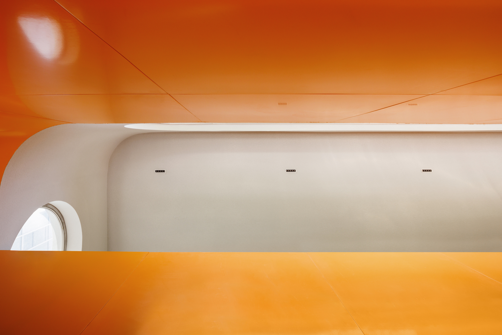 Interior view of curved white wall meeting orange ceiling planes with circular window opening
