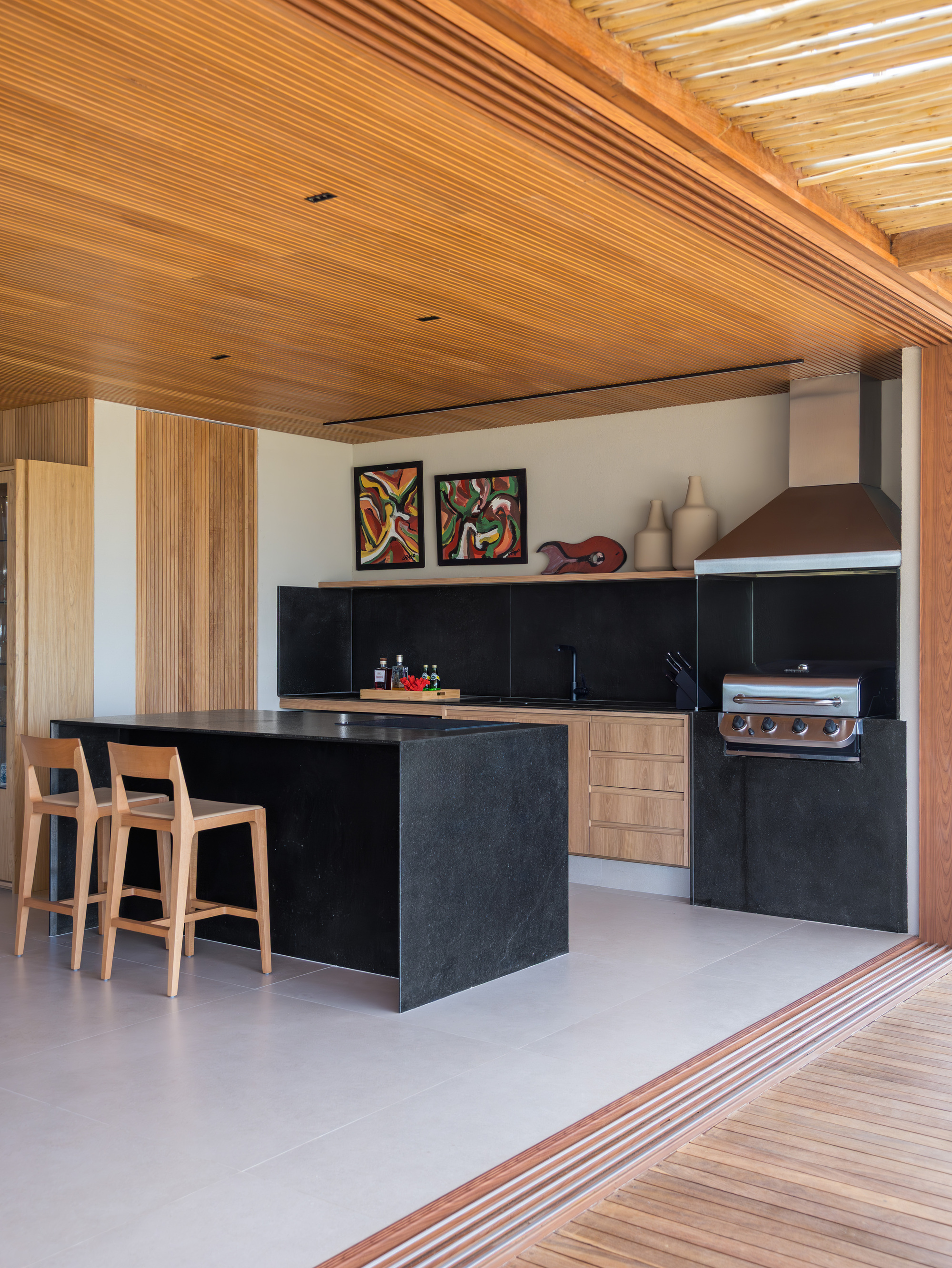 Kitchen island clad in black stone beneath an exposed timber beam and slatted ceiling opening to deck