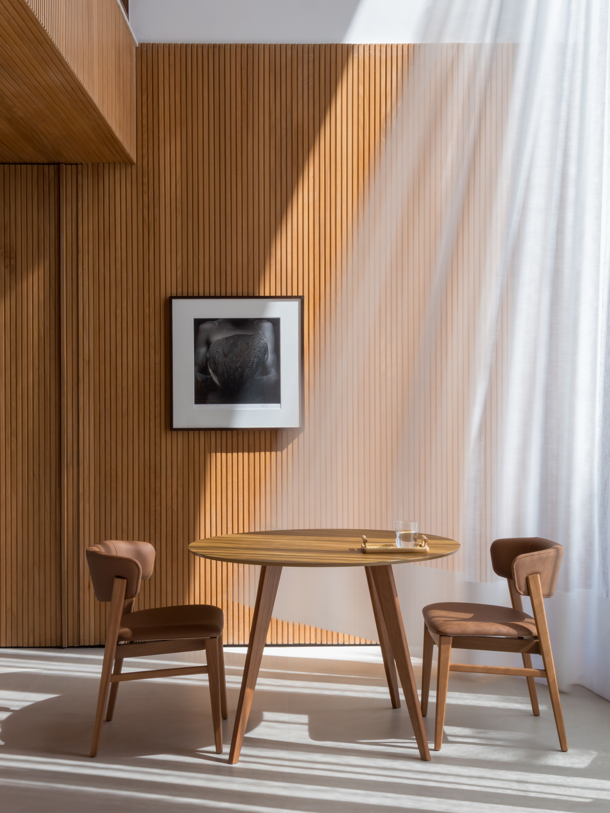 Dining area with round timber table and two chairs against slatted wood wall in afternoon light