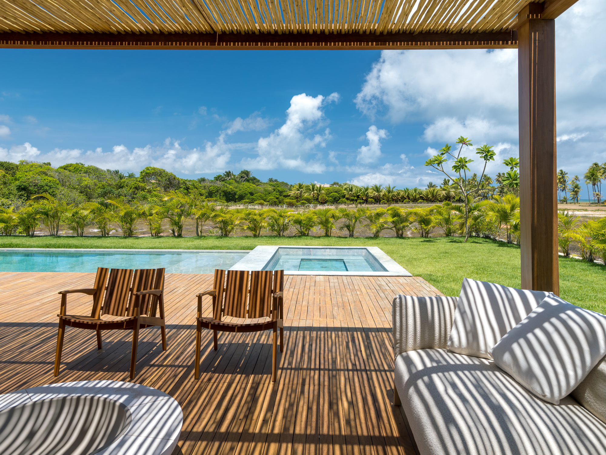 Covered terrace with slatted timber pergola overlooking pool deck and lawn under blue sky