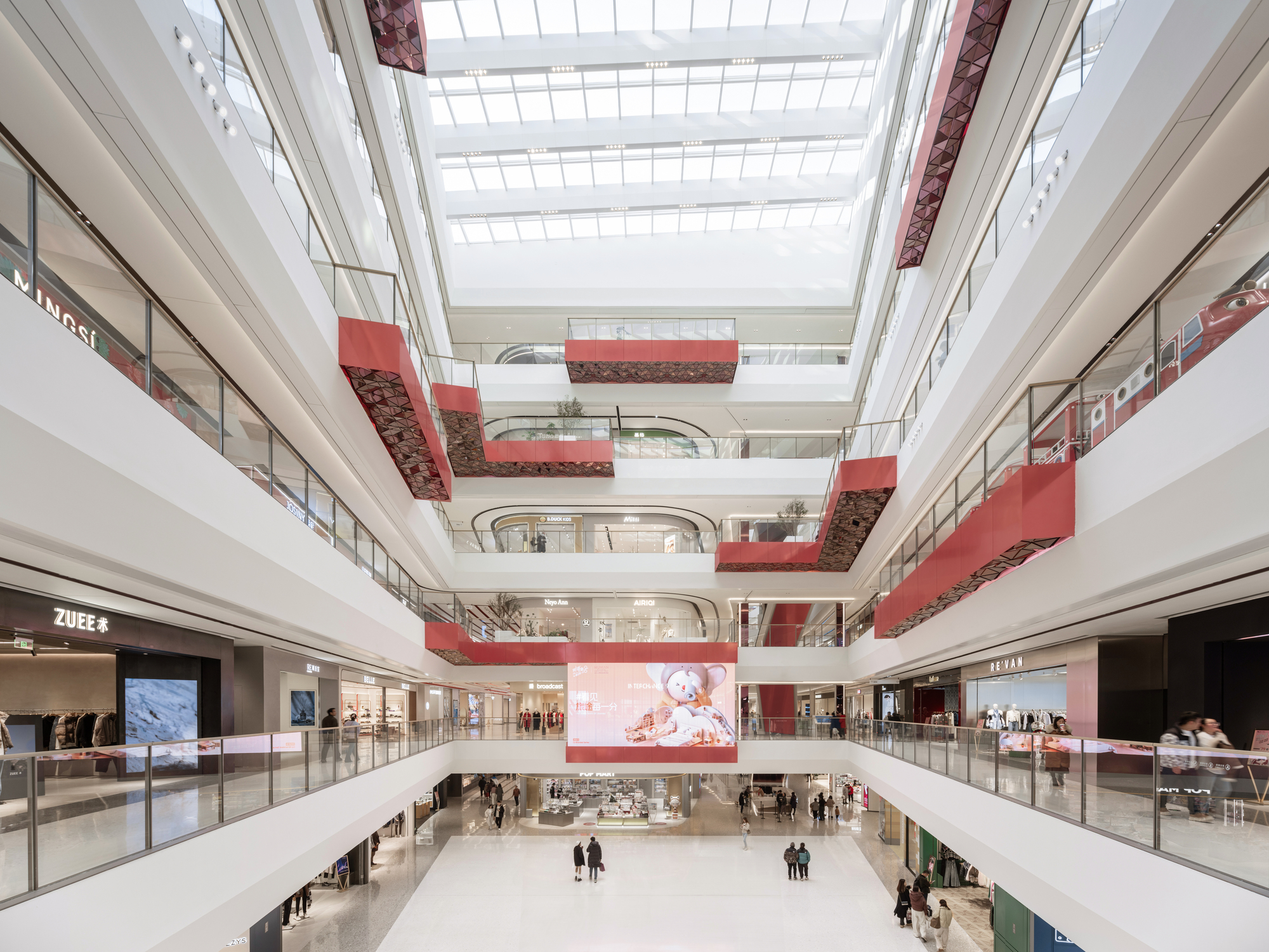 Multi-level atrium with curved balconies clad in metallic red tiles beneath a gridded skylight