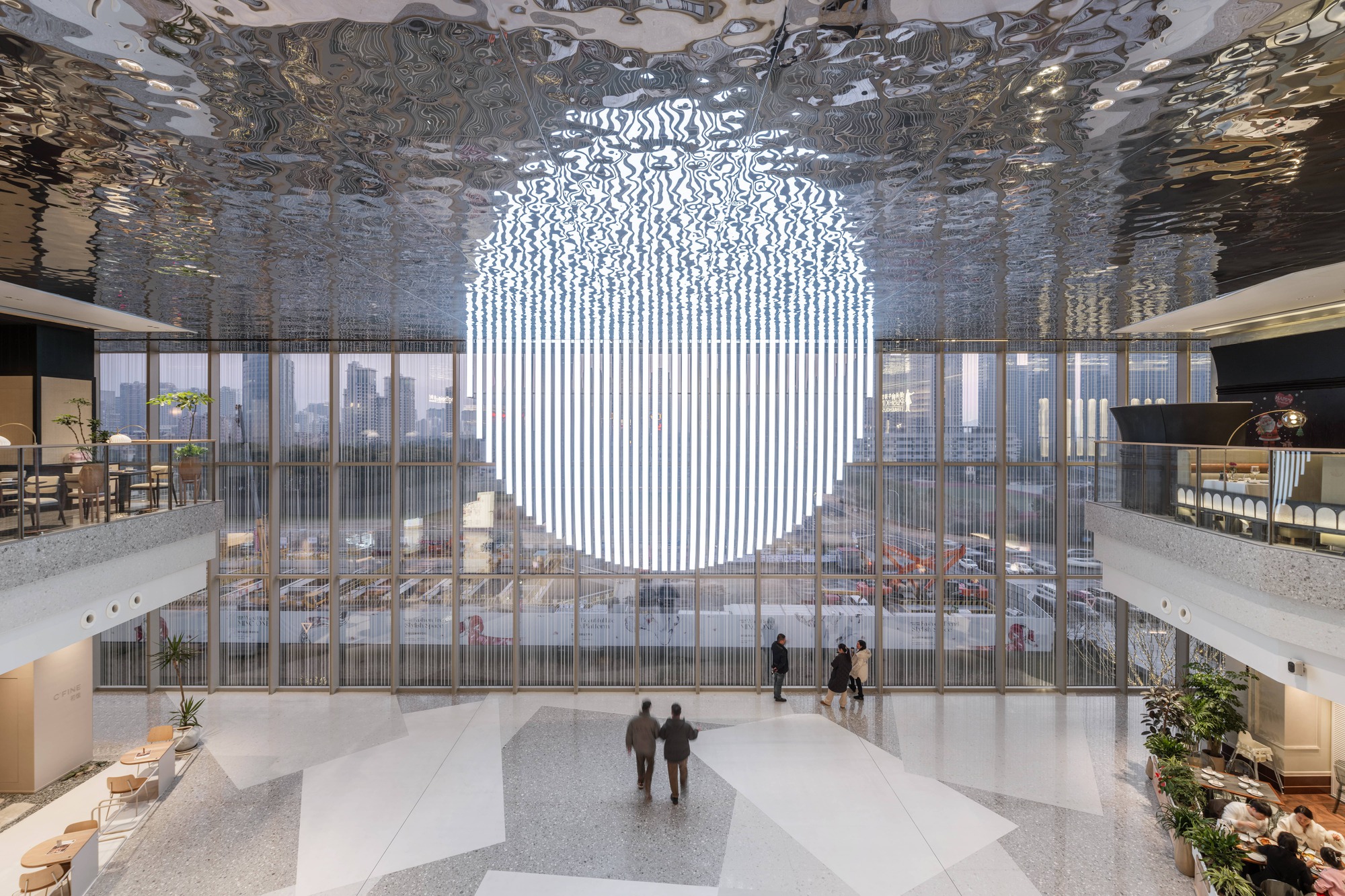 Multi-story atrium with reflective ceiling, suspended light installation, and terrazzo floors with visitors below