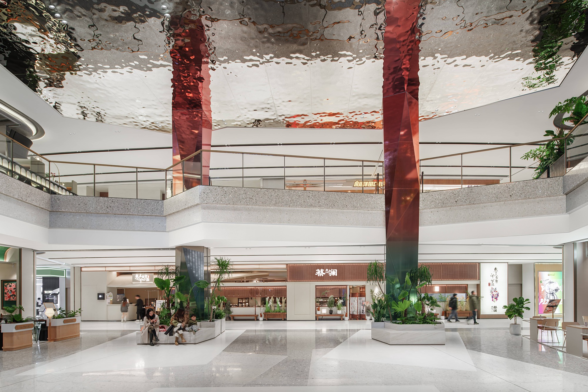 Interior mall concourse with red wrapped columns, reflective ceiling, and planters between retail storefronts