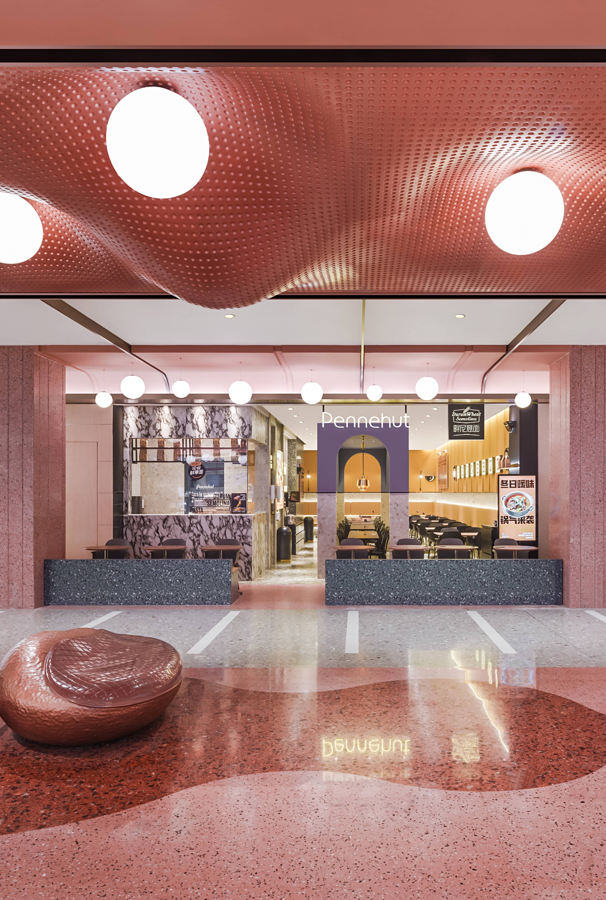 Mall seating area with undulating red perforated ceiling, terrazzo floor inlay, and restaurant frontages beyond