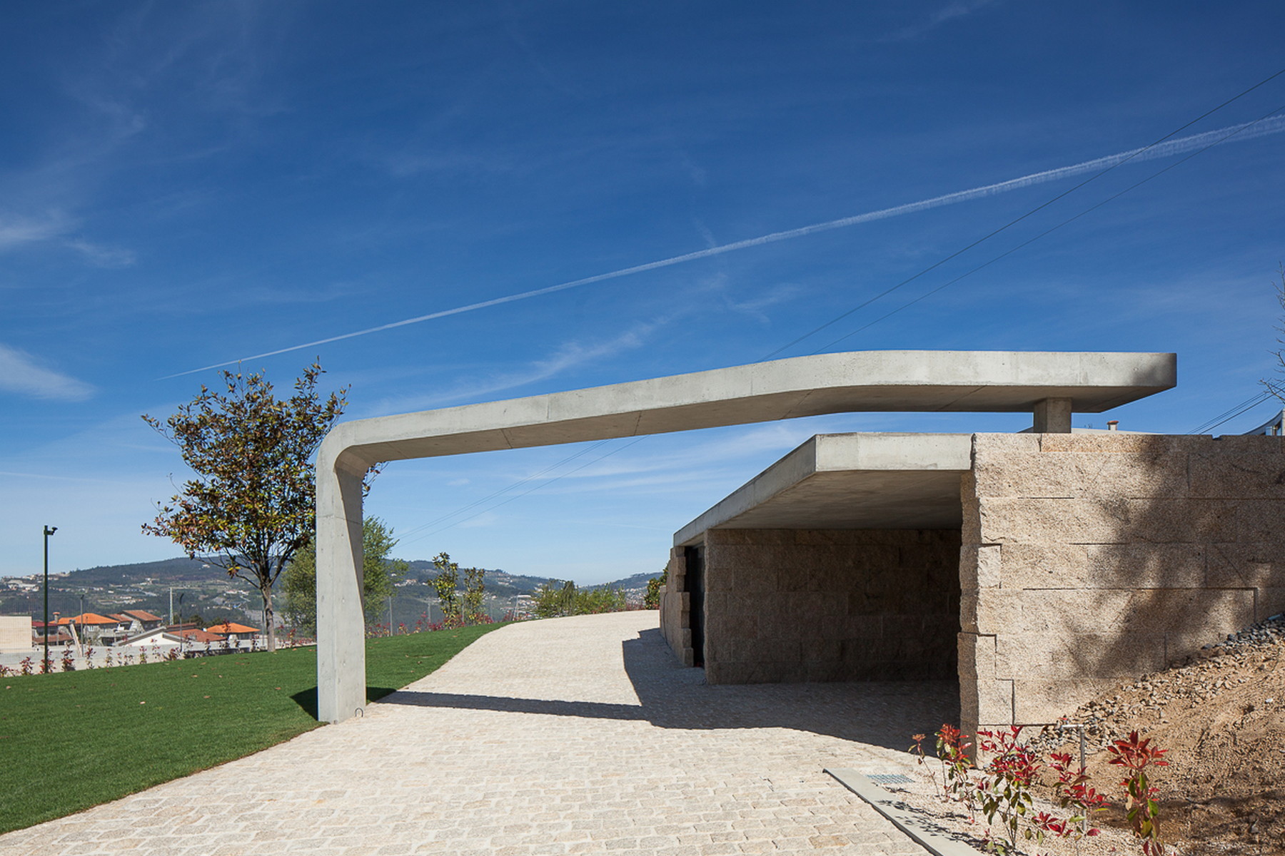 Thin concrete canopy arching over a granite-walled entrance on a cobbled path, with a jet contrail in the sky