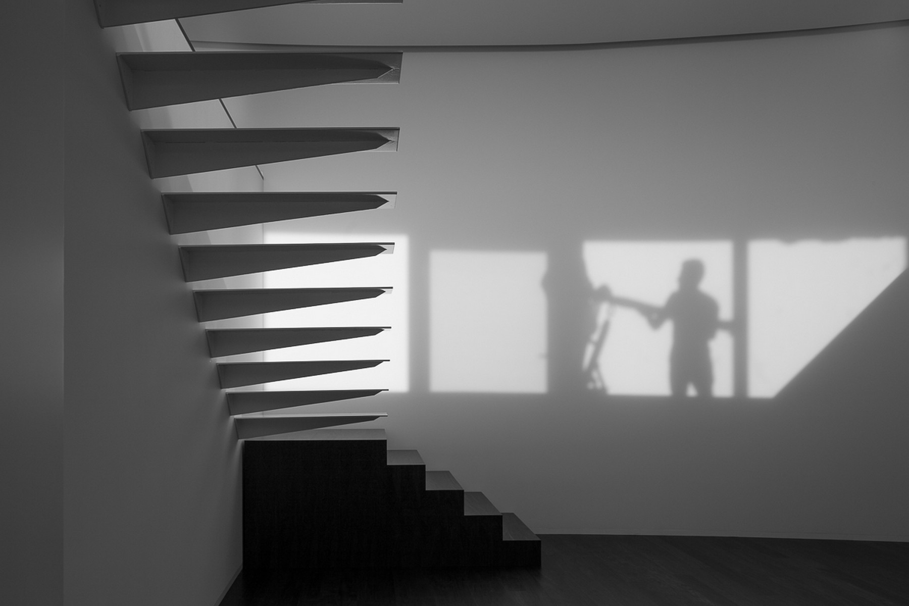 Black and white view of a cantilevered stair with tapered treads, a figure's shadow cast on the wall by window light