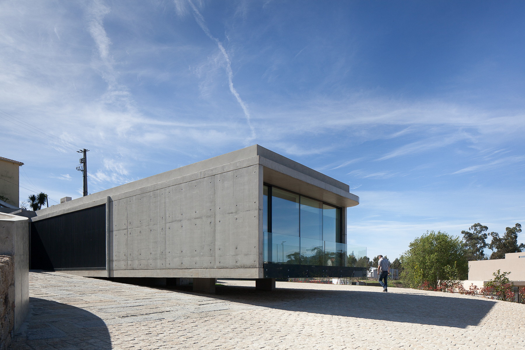 Rectangular board-marked concrete volume with a glazed end, raised over a granite cobbled forecourt as a pedestrian walks past