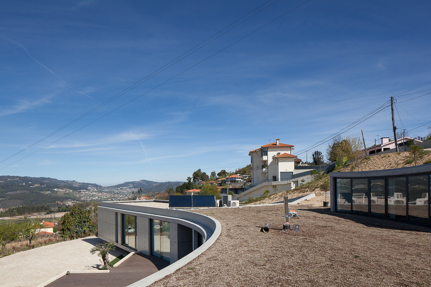 Curved green-roof wings with glazed walls looking over a hillside village, with solar panels and gravel in the foreground
