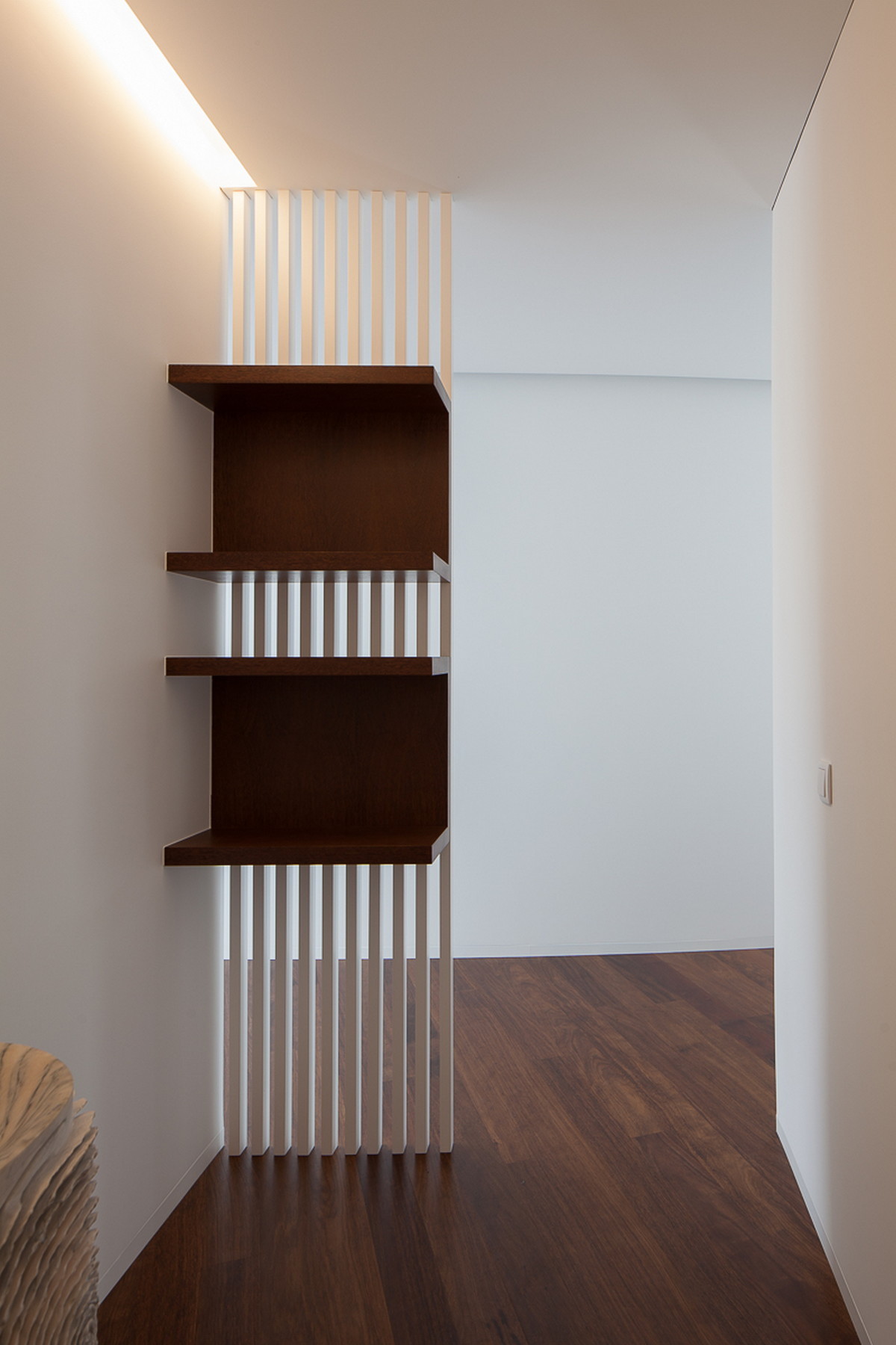 Dark timber shelving set into a white vertical slat screen, lit by a concealed cove light above the hallway