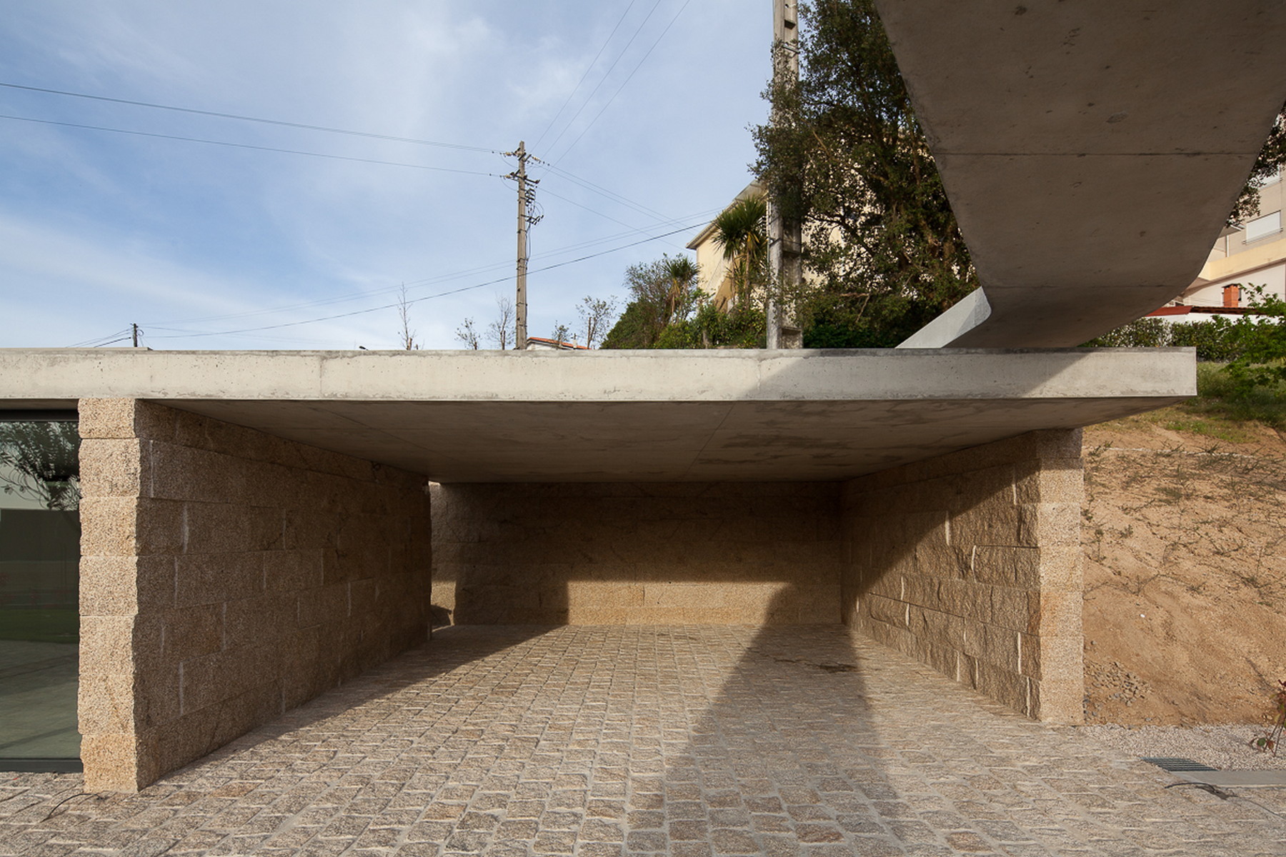 Open carport under a concrete slab with granite block walls and cobbled paving, beneath a pale blue sky