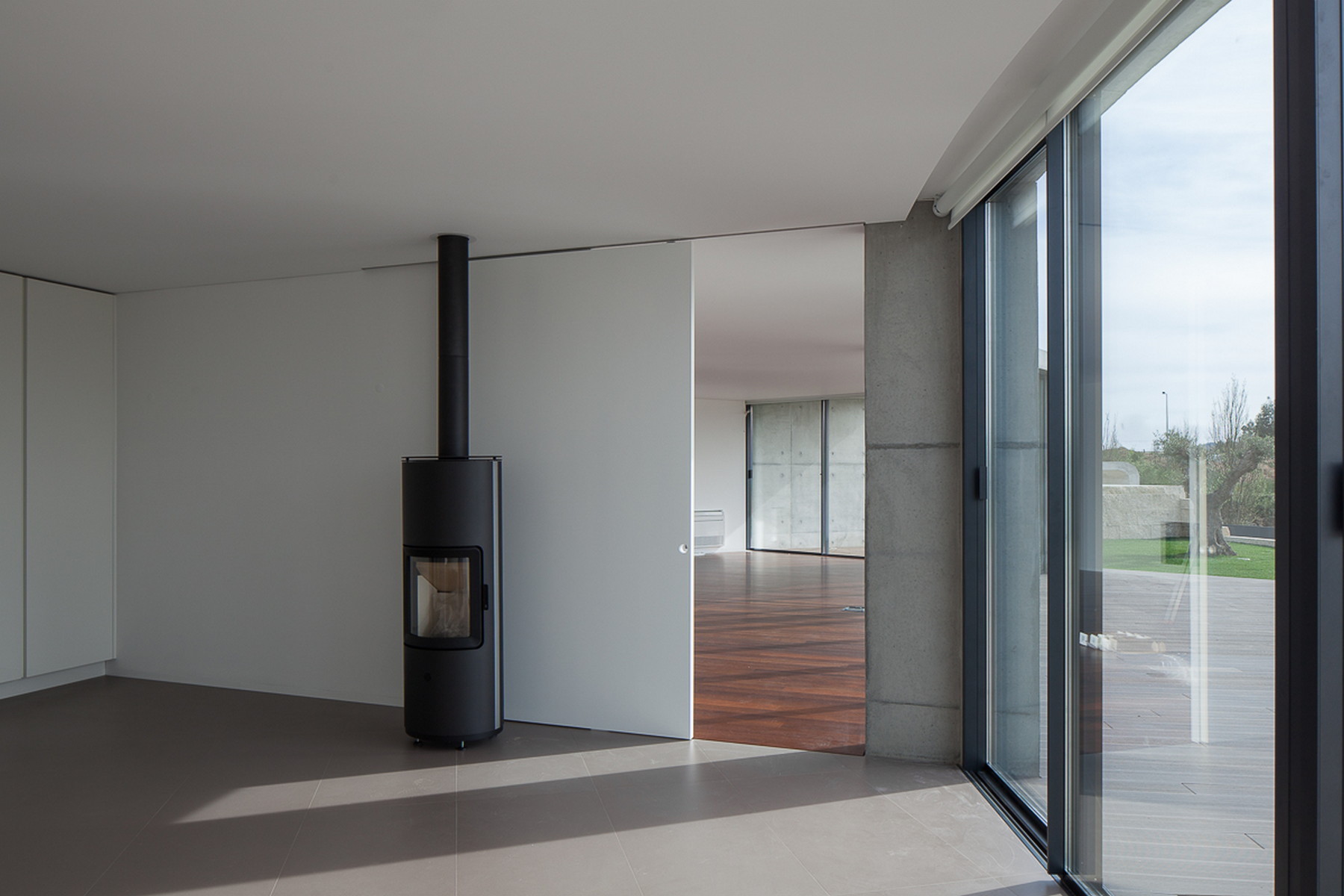 Black freestanding wood stove beside a tall white sliding door, with sunlight falling across the grey tile floor