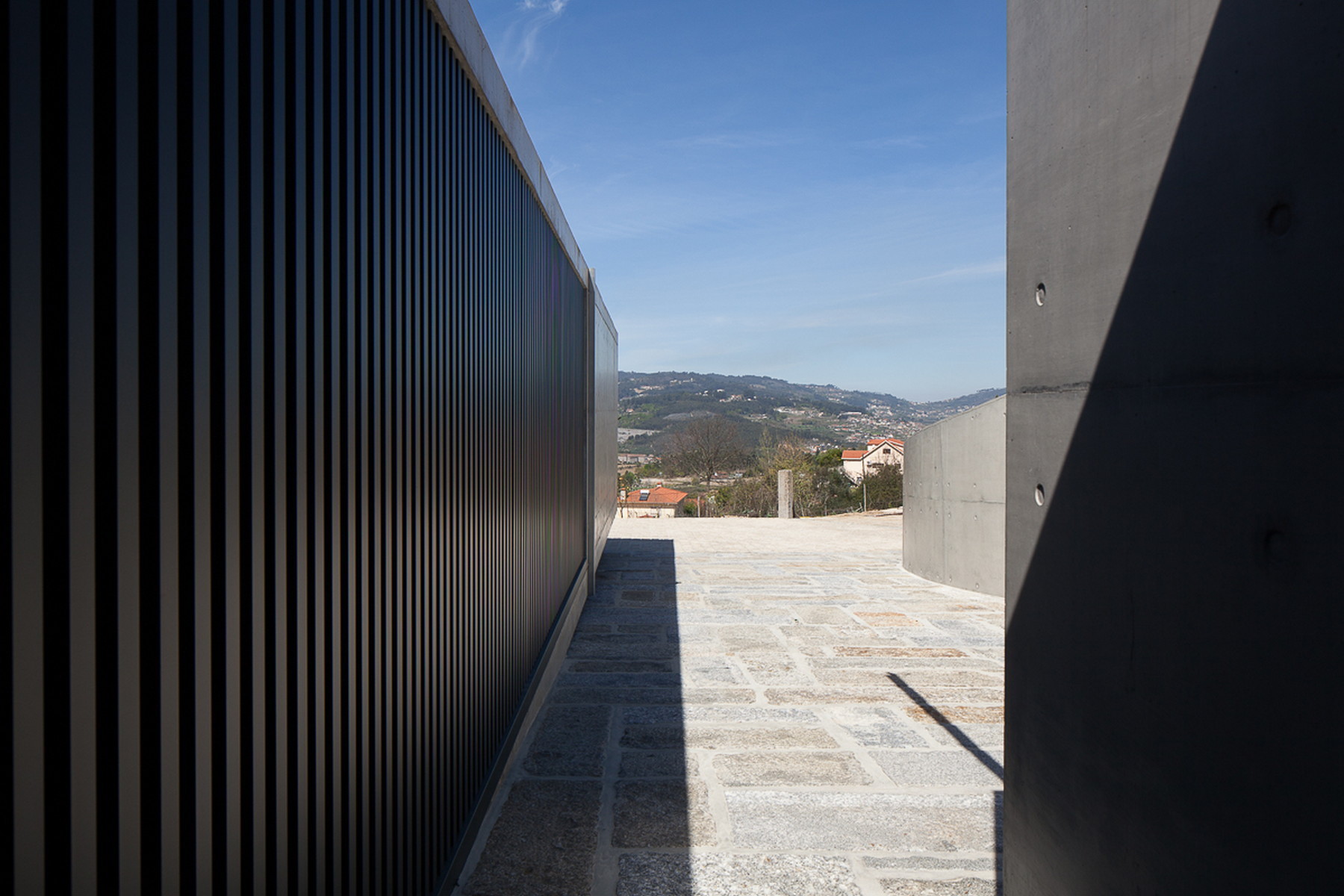 Narrow granite-paved passage between a black slatted screen and a board-marked concrete wall, opening to hills in clear sun