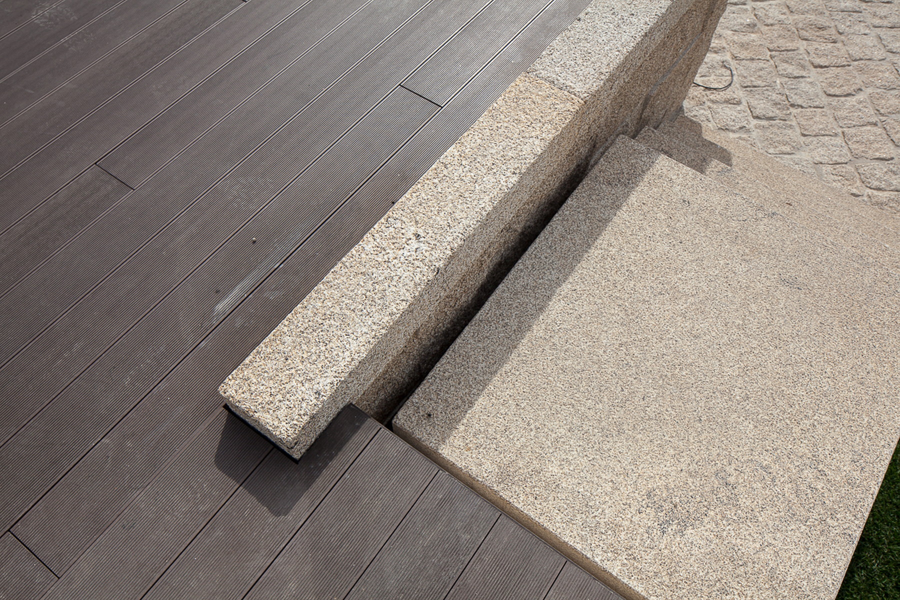 Top-down detail of a granite curb and step meeting grey composite decking, with cobbles at upper right in hard light