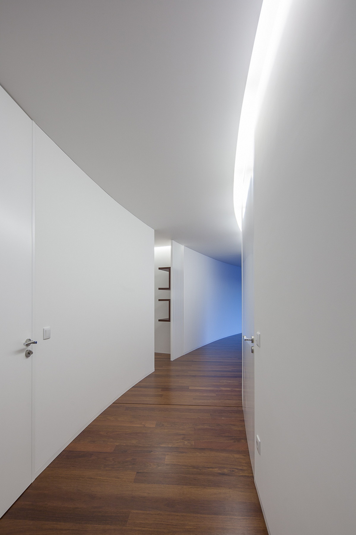 Curving white corridor with flush doors, a glowing light slot along the ceiling and a dark timber floor