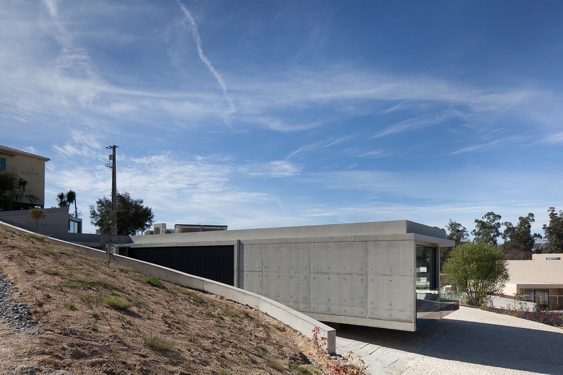 Board-marked concrete volume with a dark slatted panel, set beside a sloping bank and cobbled forecourt under streaked clouds