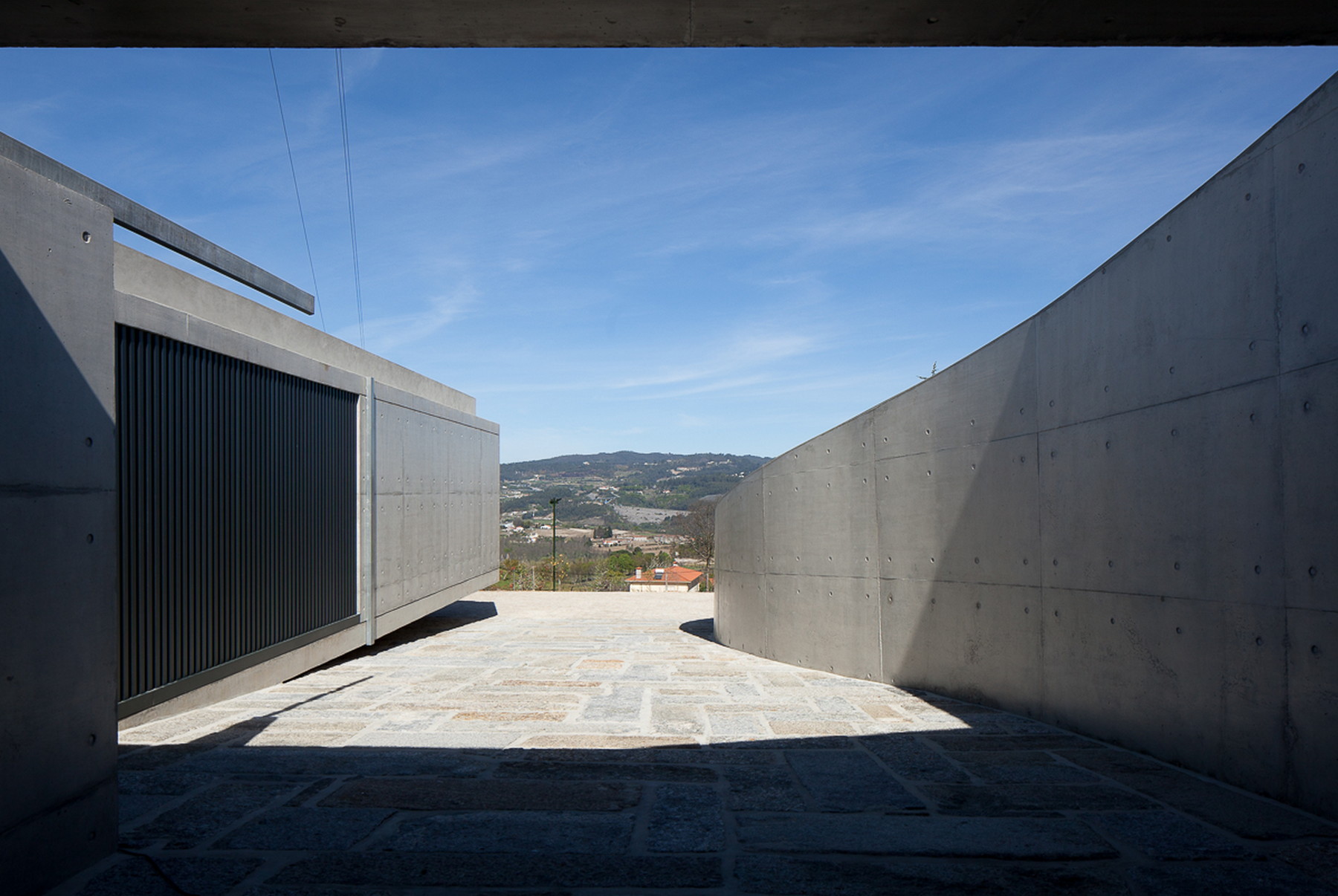 Paved granite passage between a curved concrete wall and a slatted metal gate, framing distant hills in strong sunlight