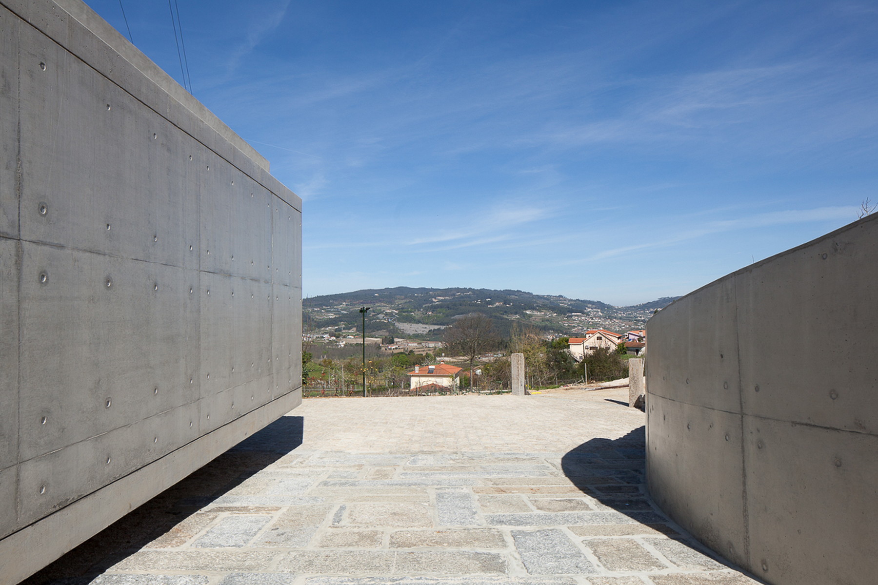 Gap between two concrete walls with tie-hole marks, opening onto a stone-paved terrace and a hazy valley view