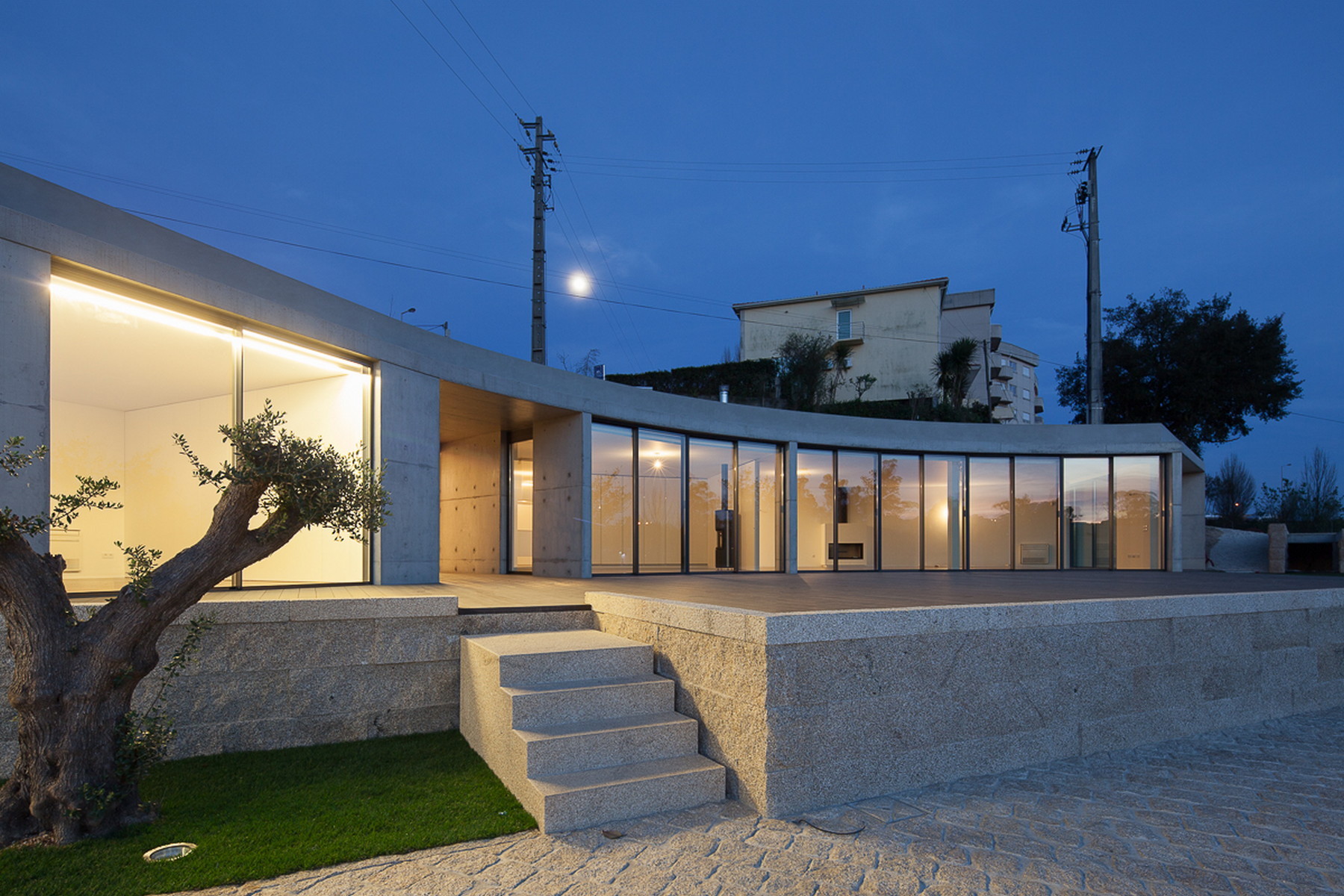 Curved glass facade glowing at dusk above a granite plinth, with an olive tree and stone steps in front