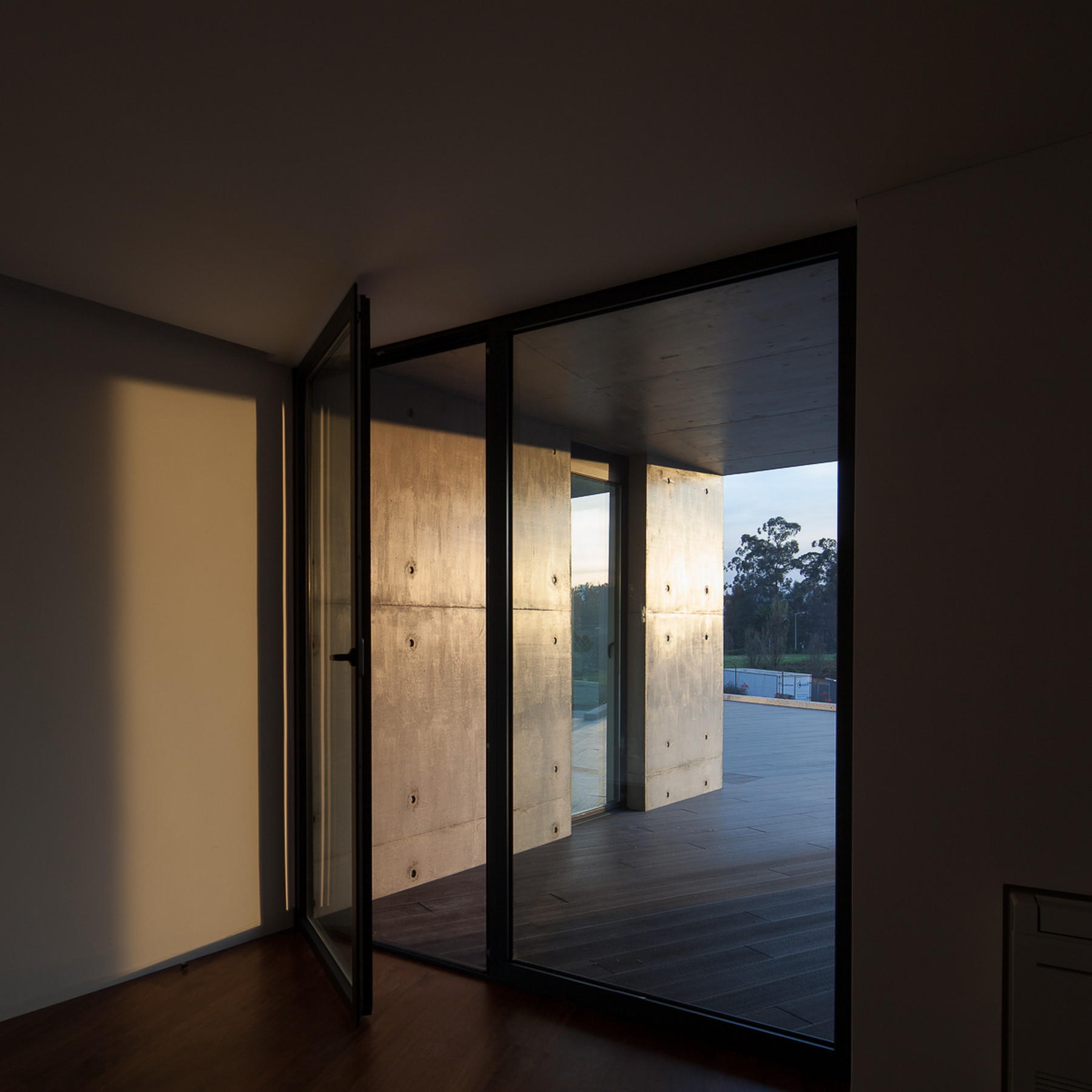 Open glazed door looking from a dark room onto a covered timber deck and concrete walls lit by low golden sun