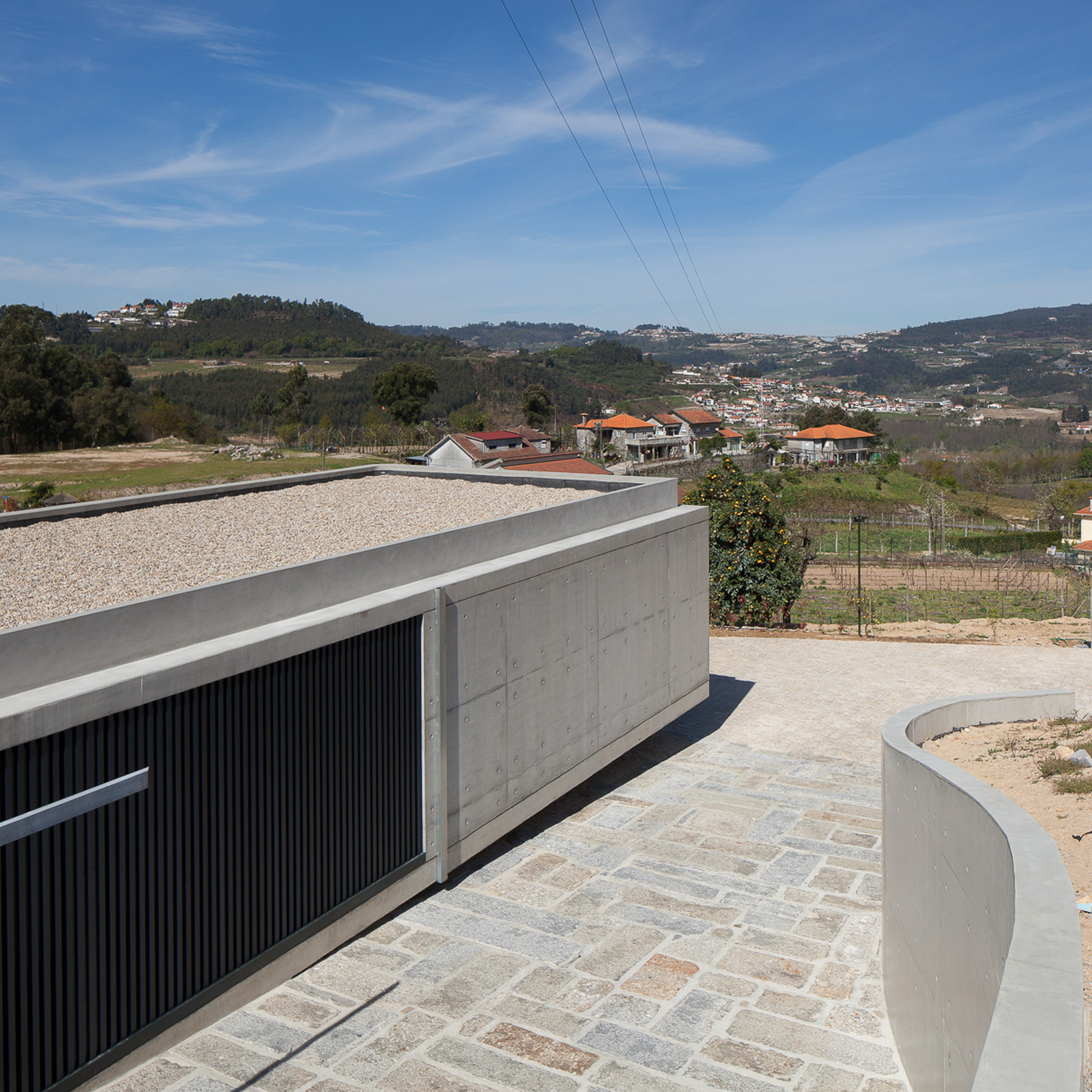 Gravel-topped concrete roof edge beside a black slatted panel and curved retaining wall, with paved stone forecourt under a blue sky
