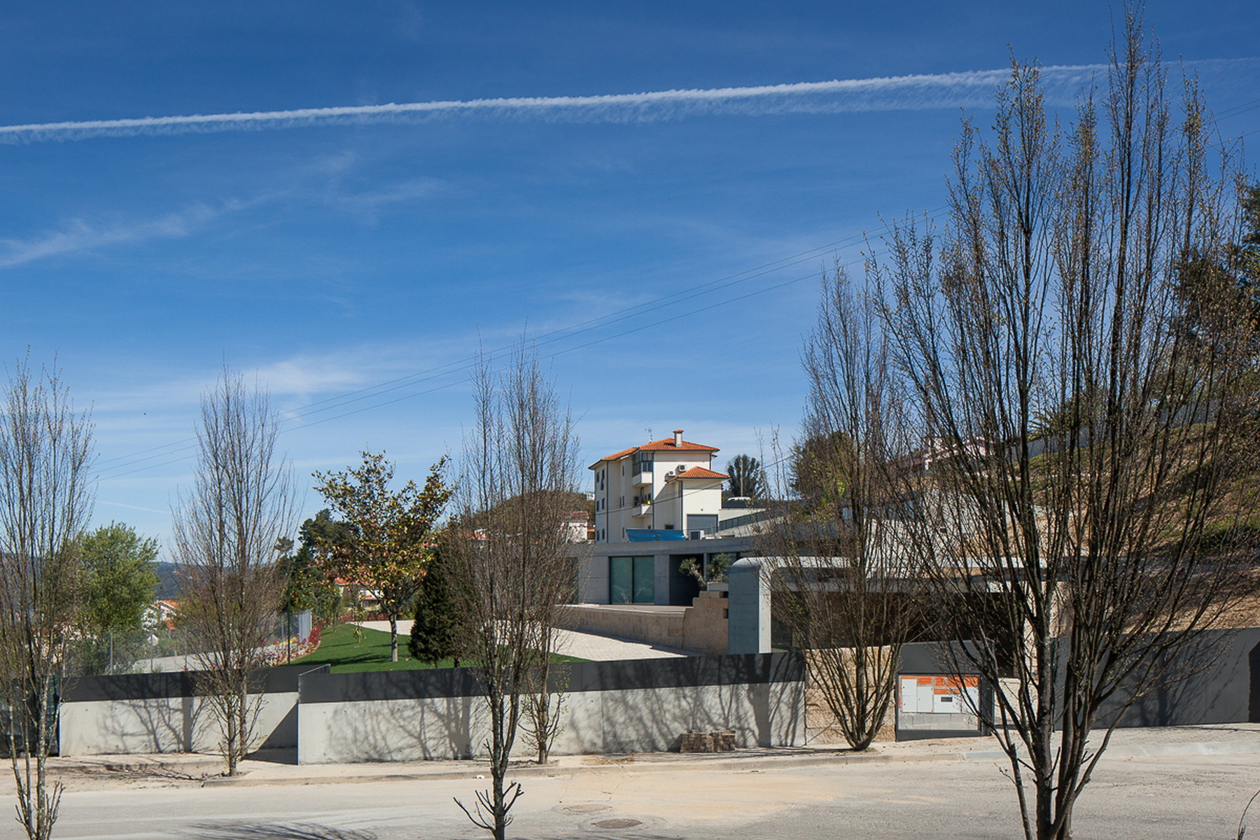 Street view of a low concrete boundary wall with the house rising behind bare winter trees and a contrail overhead