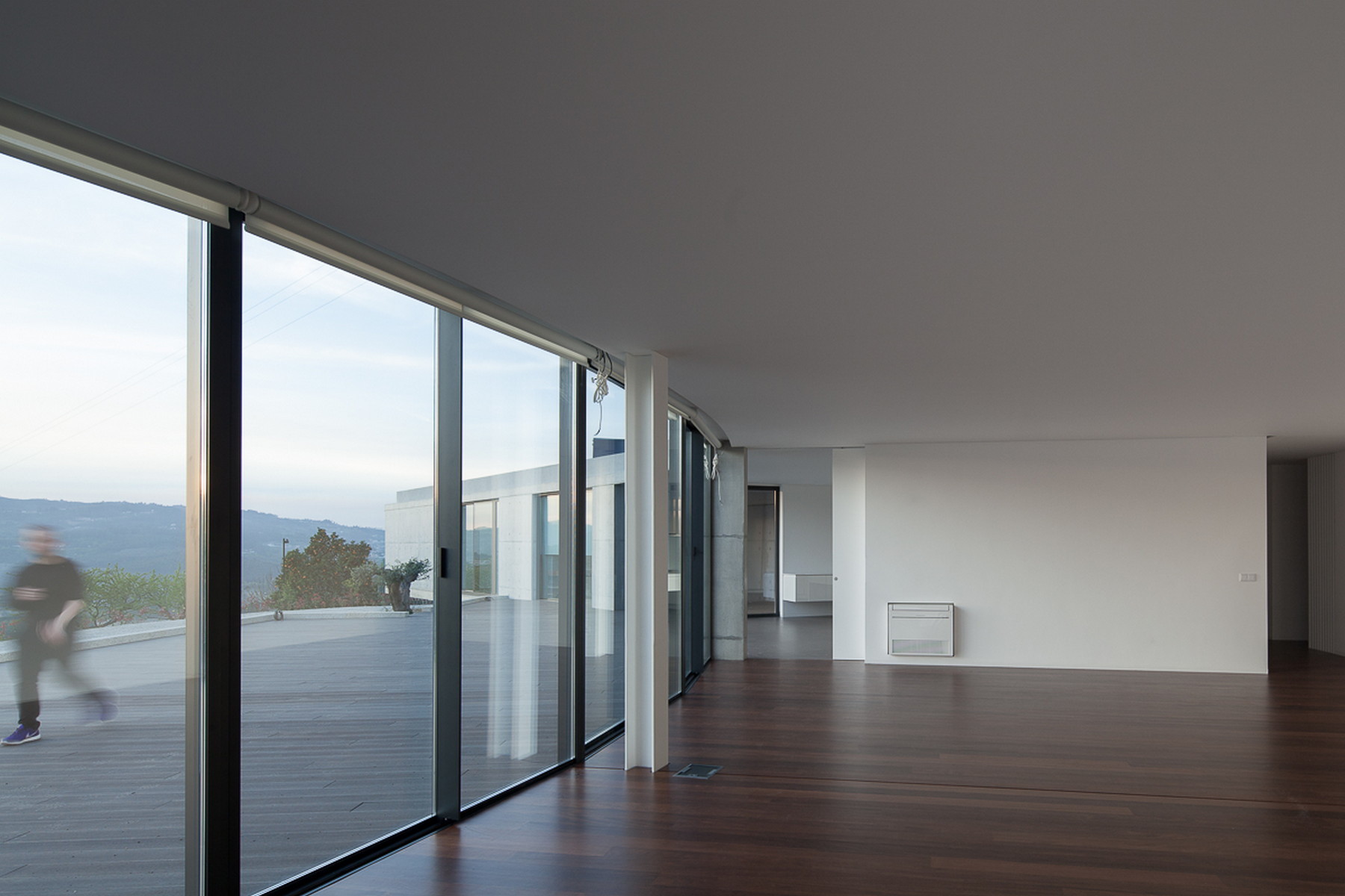 Open living room with floor-to-ceiling sliding glass and dark timber floor, a blurred figure walking across the deck outside