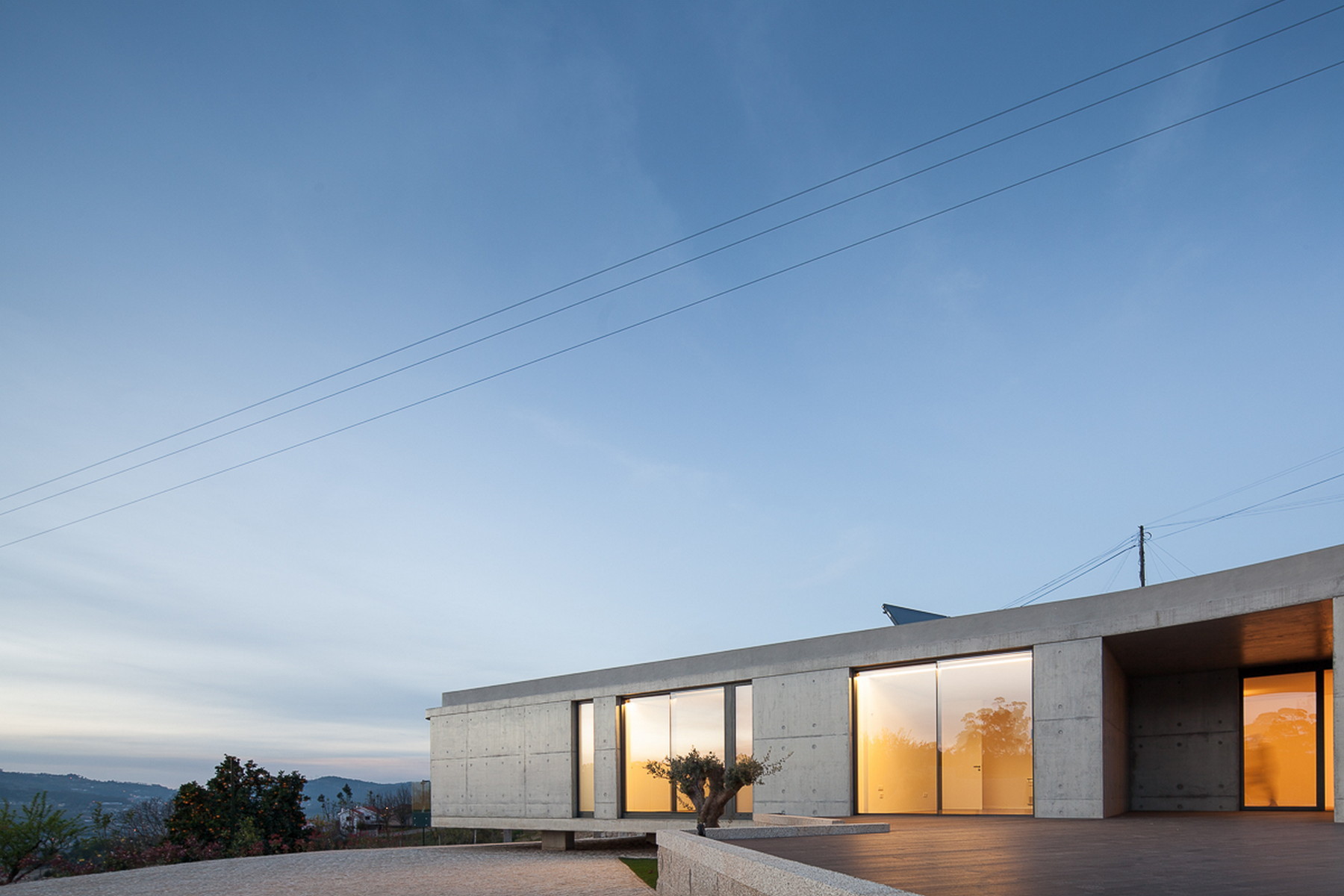 Board-formed concrete house with glowing sliding glass doors and a covered timber deck beneath a dusk sky crossed by power lines
