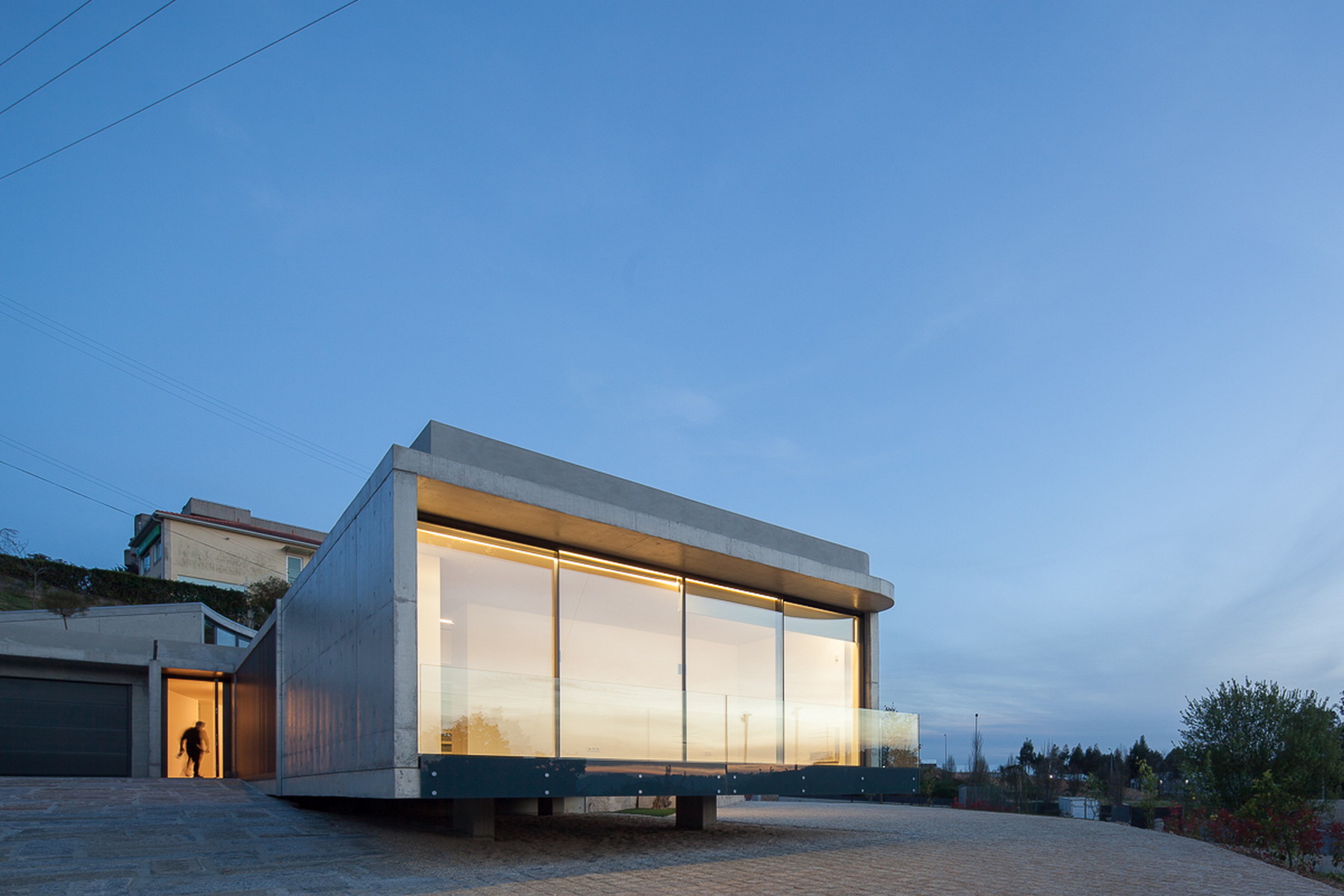 Cantilevered concrete volume with curved glass corner lit from within, a figure standing in the entry doorway at dusk