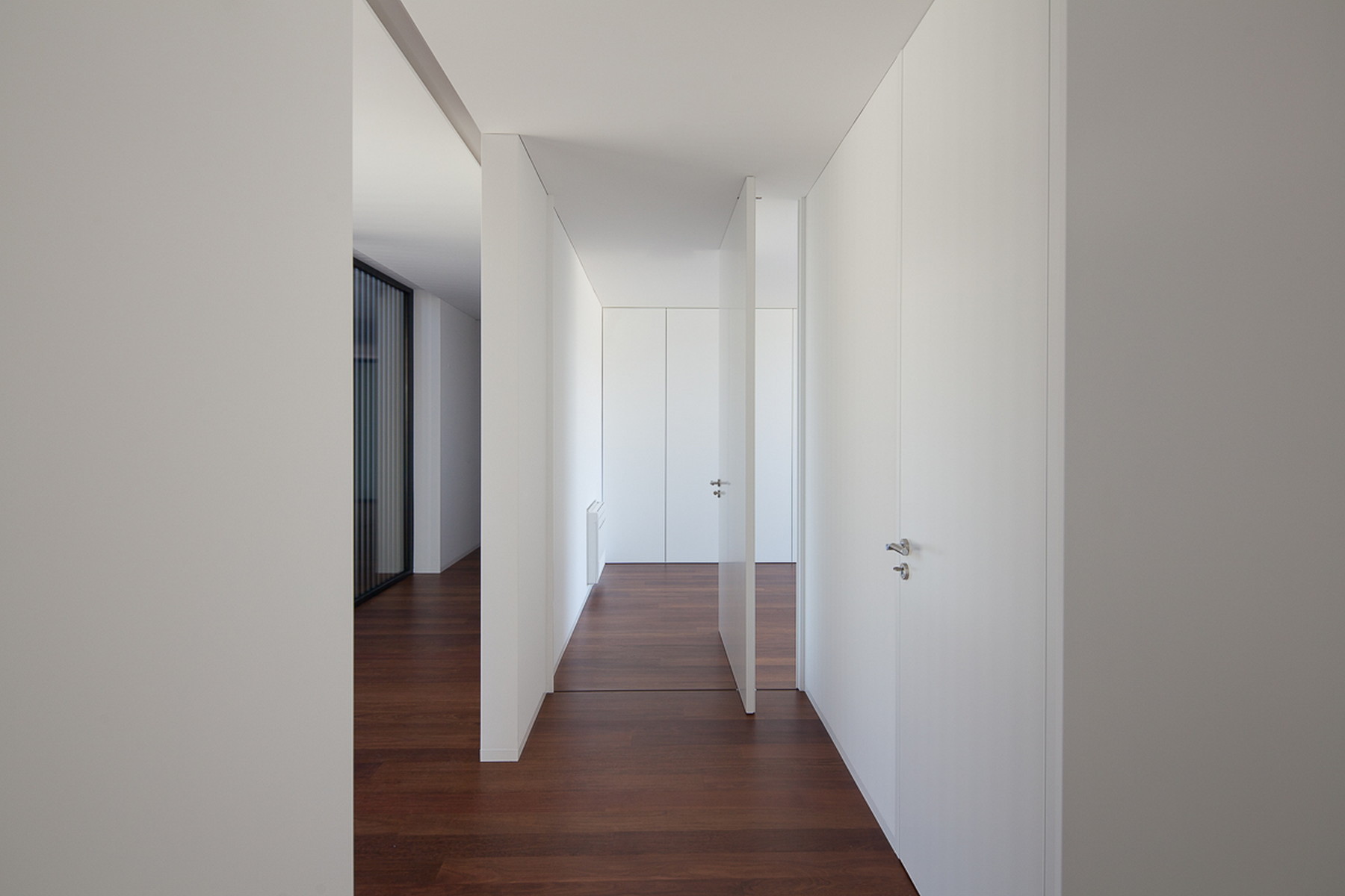 Narrow corridor of flush white doors and panels with a pivoting door ajar over dark wood flooring