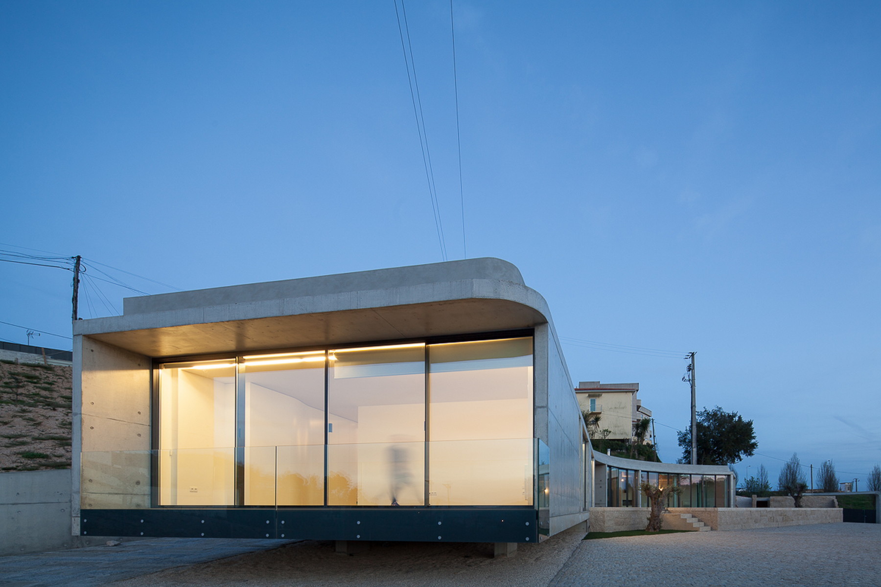 Glass-walled concrete pavilion with rounded roof edge raised on thin supports, glowing at blue hour beside a gravel court
